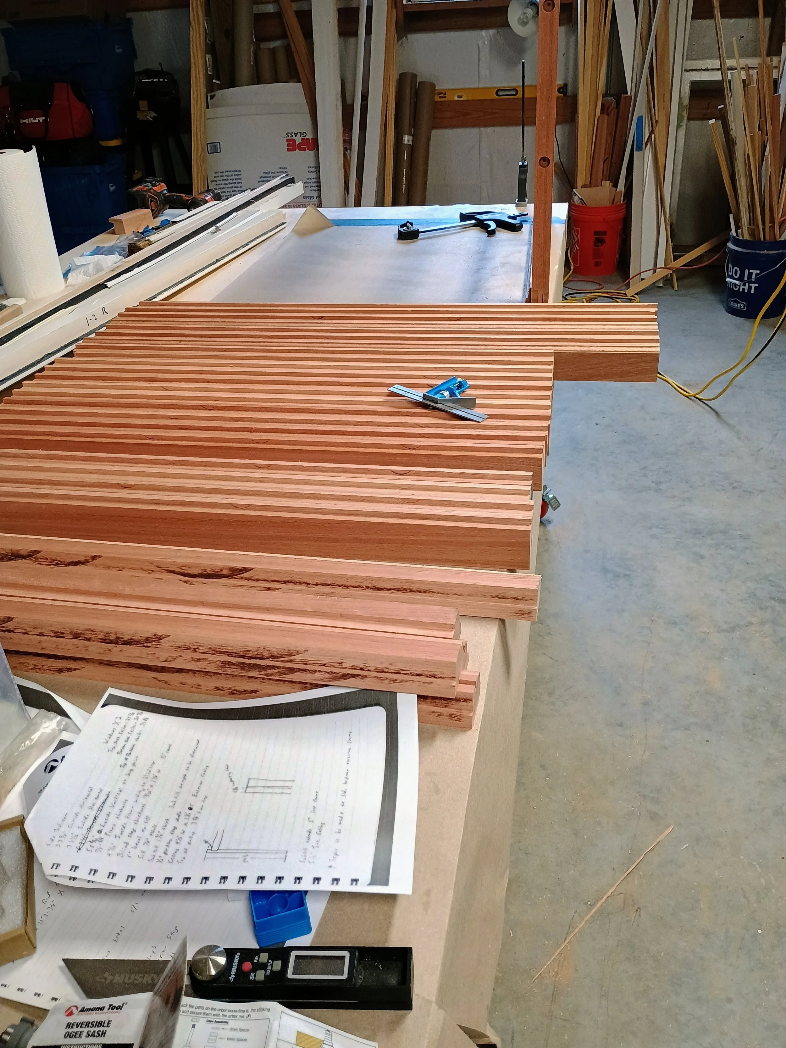 Woodworking workshop with stacks of wooden slats on the workbench, measuring tools, handwritten plans, and various tools and equipment in background.