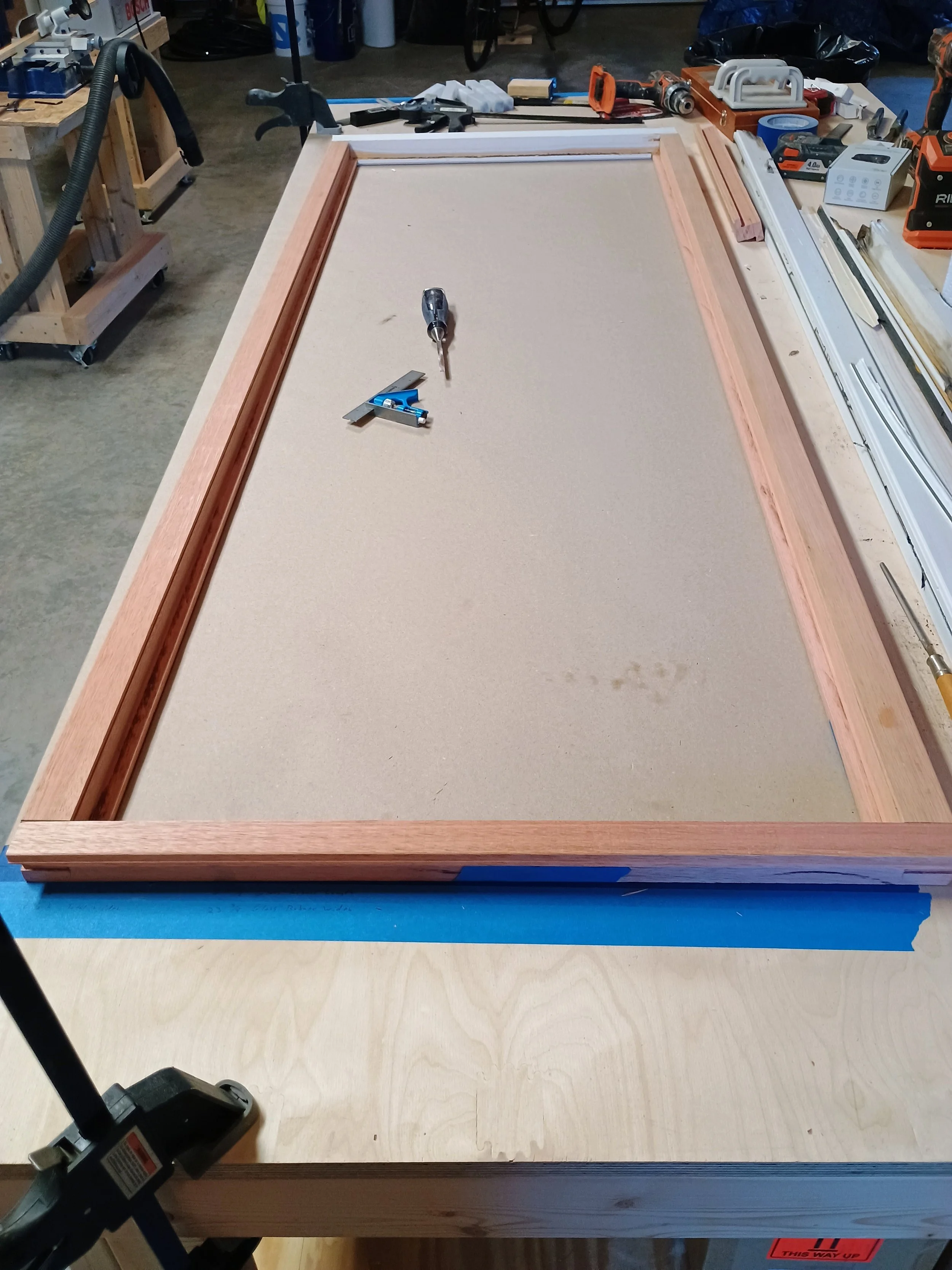 Workshop scene with a large wooden frame on a workbench, various tools, and equipment around, including a screwdriver, square, and power tools.