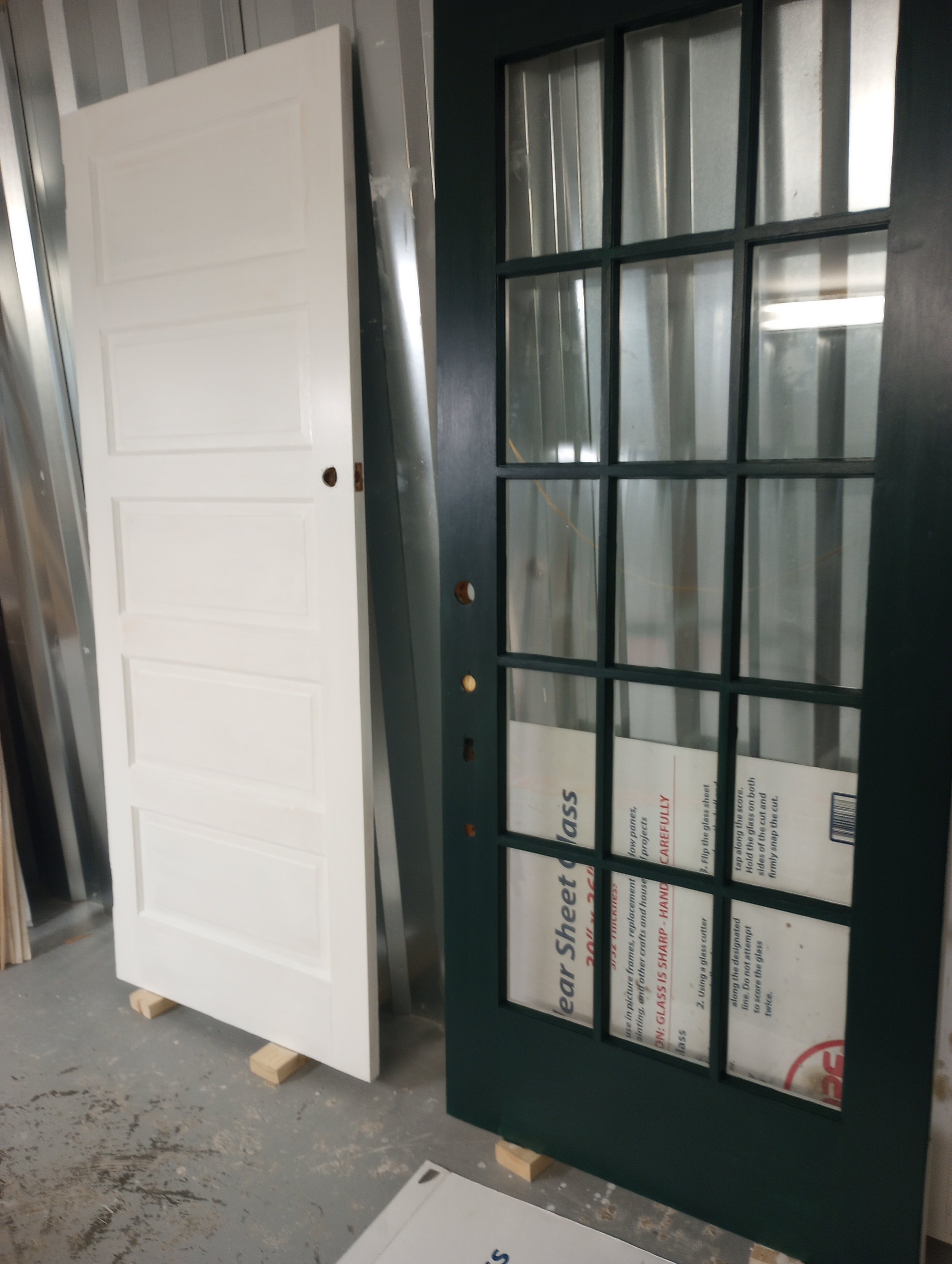 A white door leaning against a metal surface and next to a large green window frame with glass panels, set on a concrete floor.