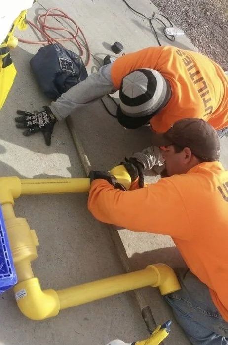 Two workers in orange shirts and gloves installing or repairing yellow underground utility pipes on a sidewalk, with tools and equipment nearby.