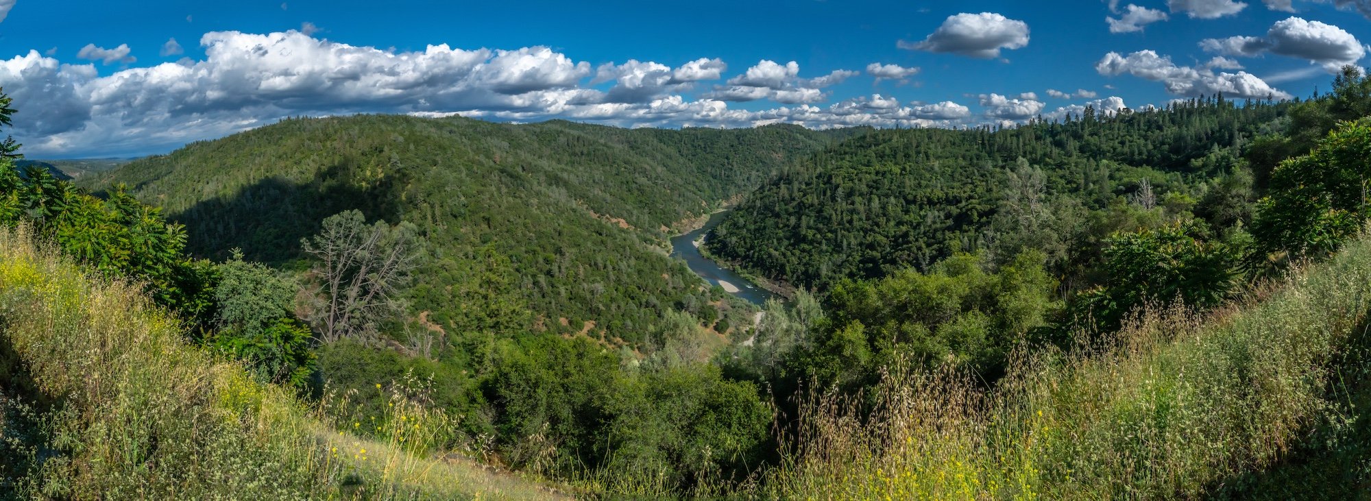 A panoramic view of a lush green valley with a winding river, surrounded by forested hills under a partly cloudy sky.