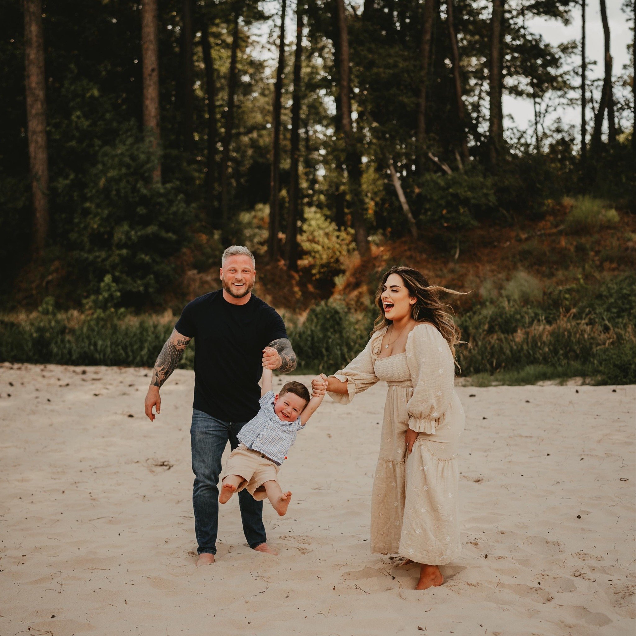 A family of three playing on the beach. The father, with tattoos and wearing a black T-shirt, is holding the hand of a young boy in a plaid shirt and khaki shorts. The mother, in a long beige dress, is also holding the boy's other hand. All three are smiling and appear to be having fun, with a forest background behind them.