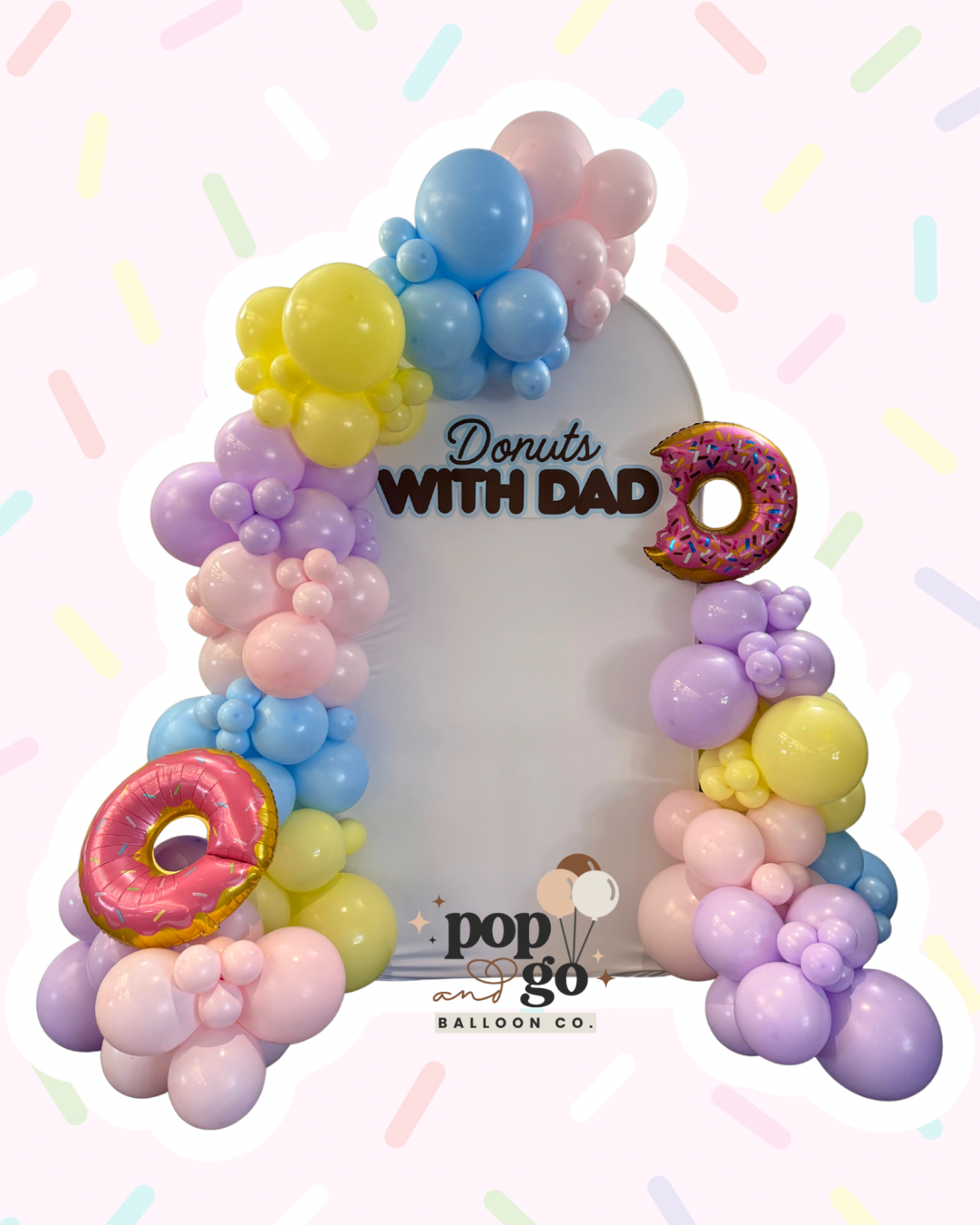 Colorful balloon arrangement with pastel pink, purple, yellow, and blue balloons, including donut-shaped balloons with sprinkles, surrounding a sign that reads "Donuts with Dad" and a balloon with "pop go" logo at the bottom.