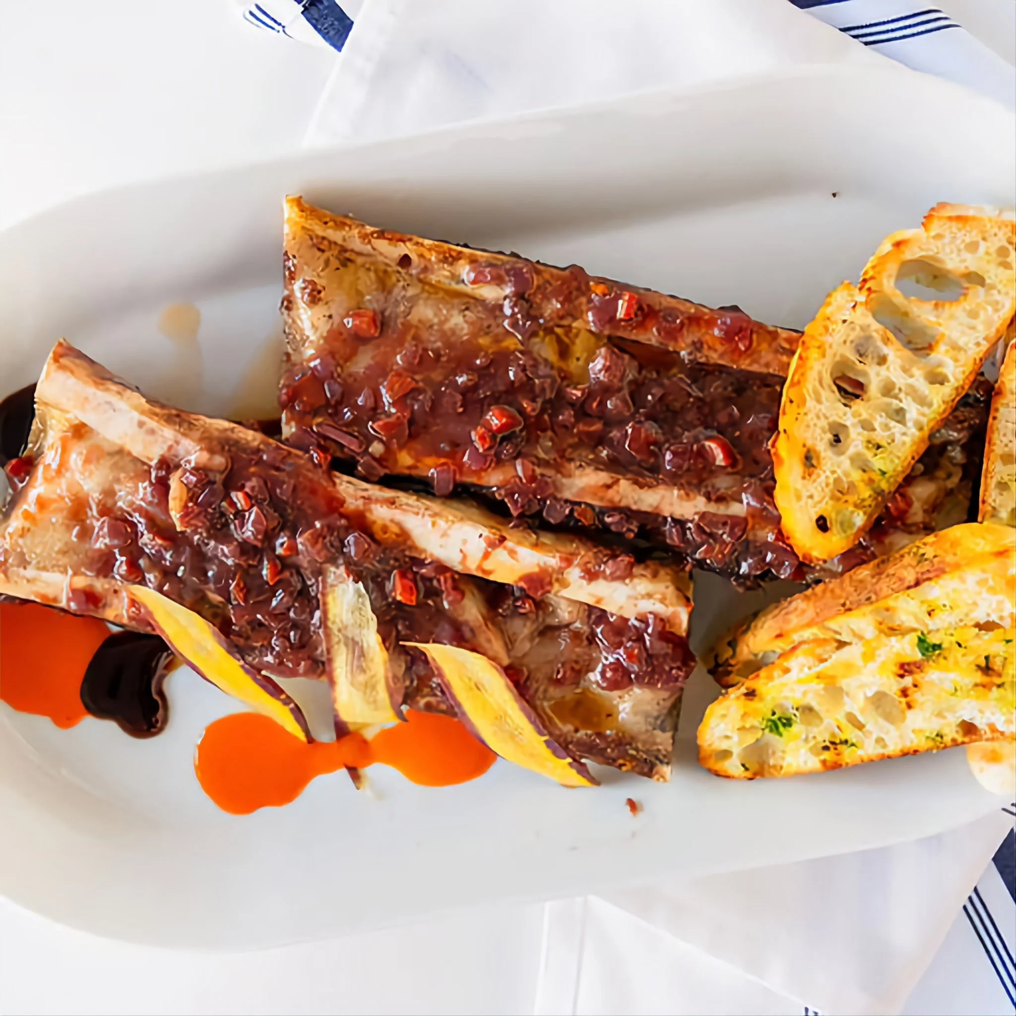 Three grilled pork ribs topped with a chili glaze, served with grilled bread slices and a drizzle of orange and balsamic sauces on a white oval plate.