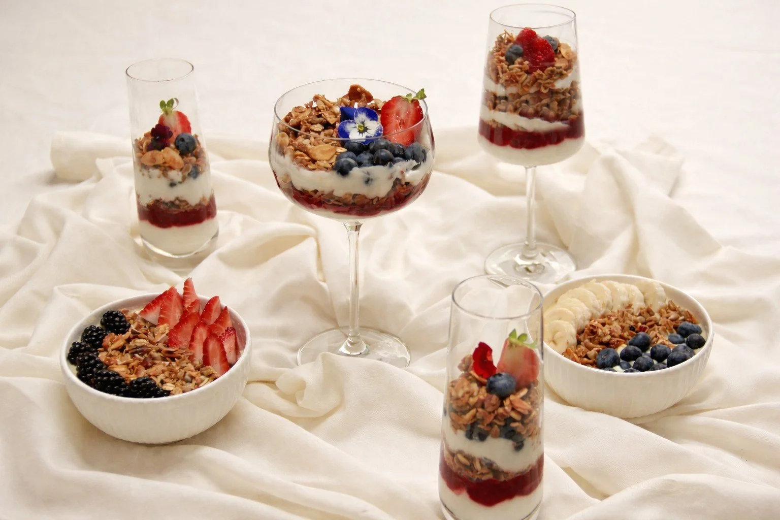 Assorted yogurt parfaits and bowls with granola, fresh strawberries, blueberries, and other berries on a white cloth background.