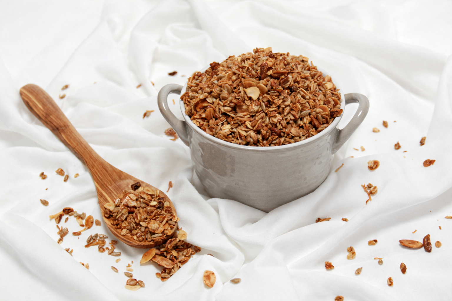 A bowl filled with chopped nuts, placed on a white cloth with scattered nuts and a wooden spoon beside it.