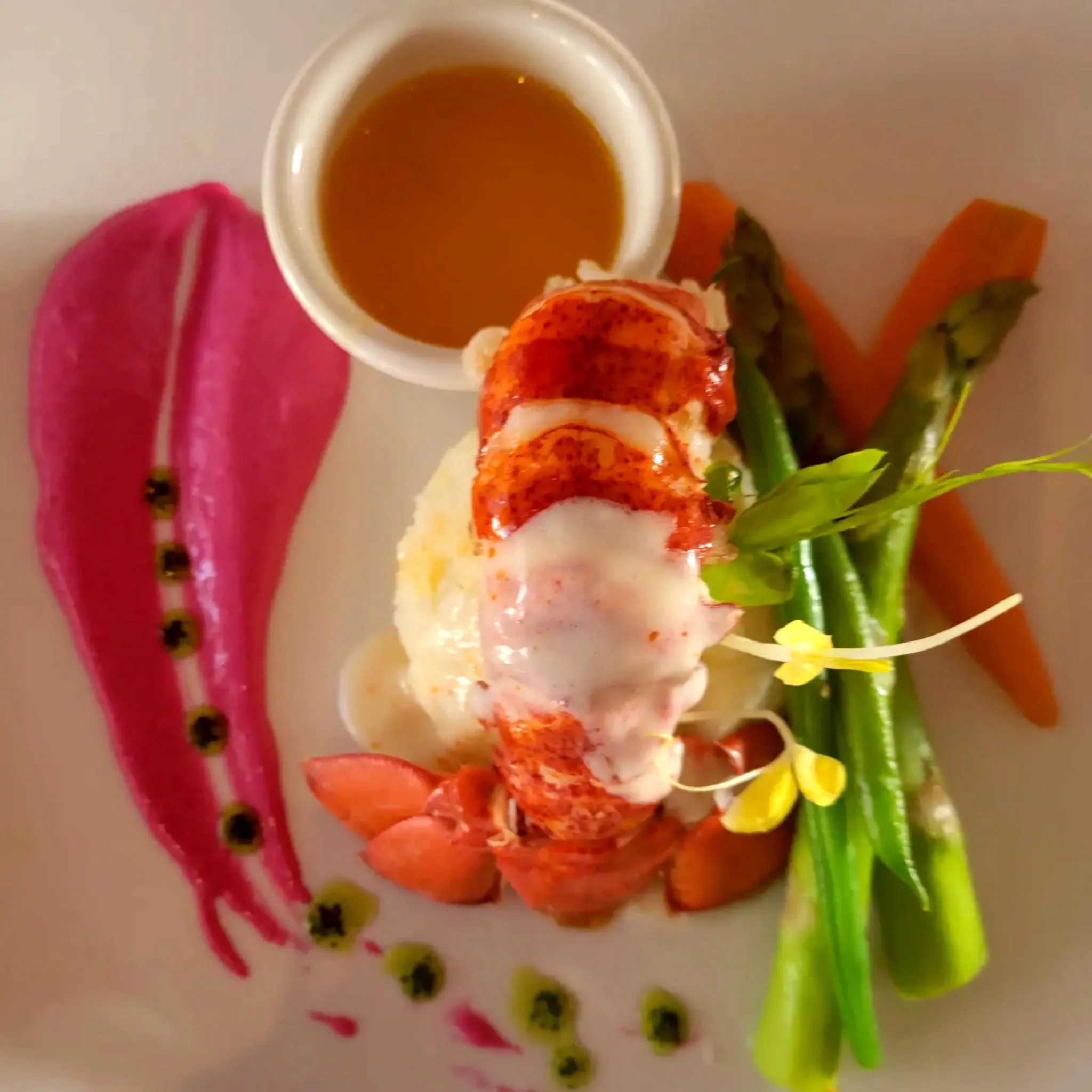 Lobster tail with melted butter, surrounded by green beans, carrots, and a pink crushed beet or berry sauce on a white plate, garnished with microgreens and edible flowers.