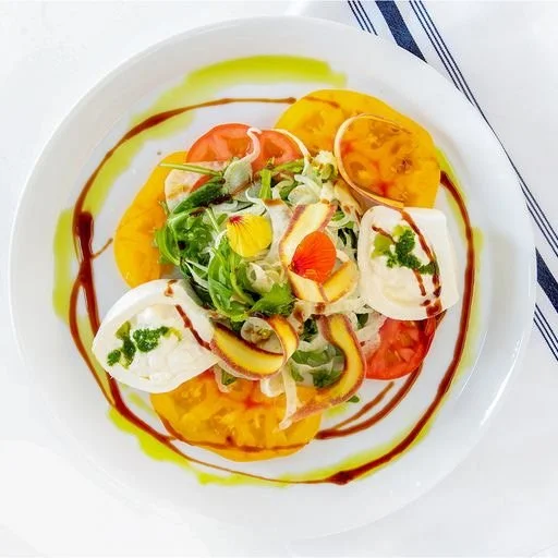 A colorful salad with mixed greens, sliced tomatoes, yellow and orange heirloom tomatoes, mozzarella cheese, and edible flowers on a white plate, drizzled with balsamic glaze and olive oil.