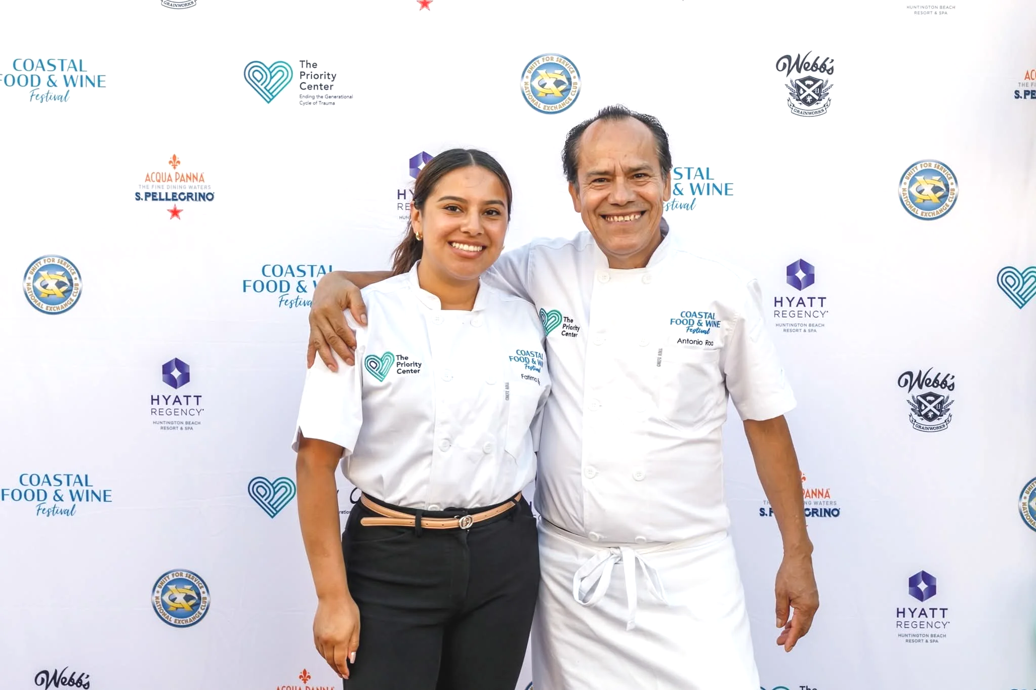 Two chefs smiling and posing together at the Coastal Food & Wine Festival, with a backdrop featuring event logos and sponsors.