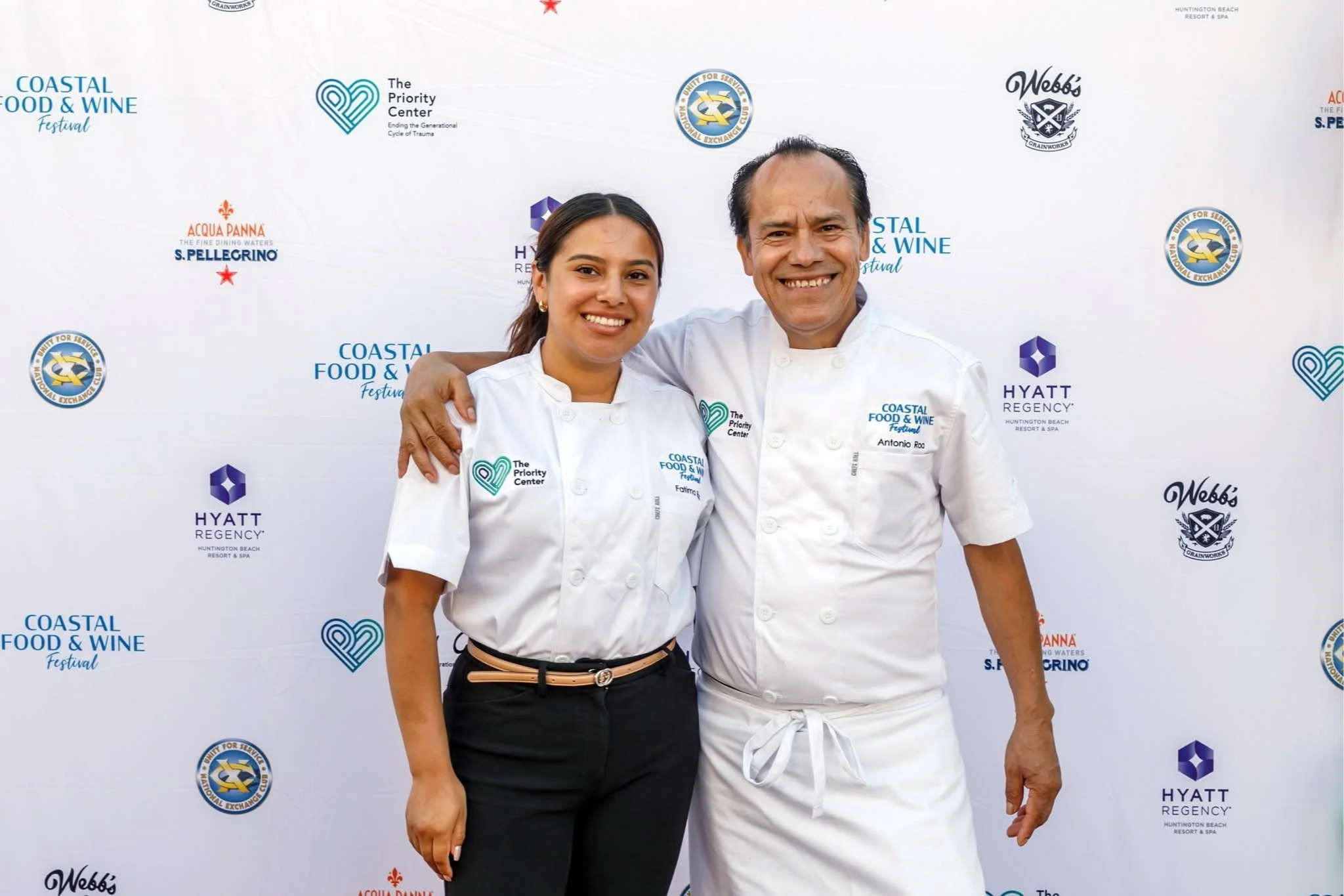 Two chefs, a woman and a man, stand together with their arms around each other, smiling at the camera, in front of a backdrop with various festival and hotel logos.