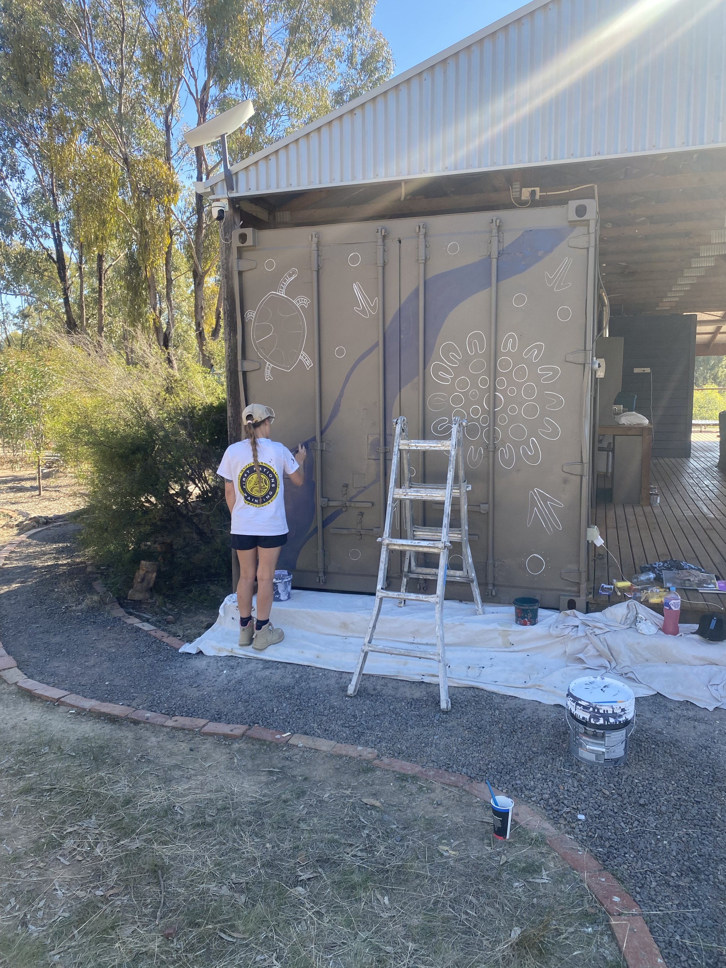 Person painting a large outdoor mural on a metal storage container, with various paint supplies on the ground, surrounded by trees and outdoor scenery.