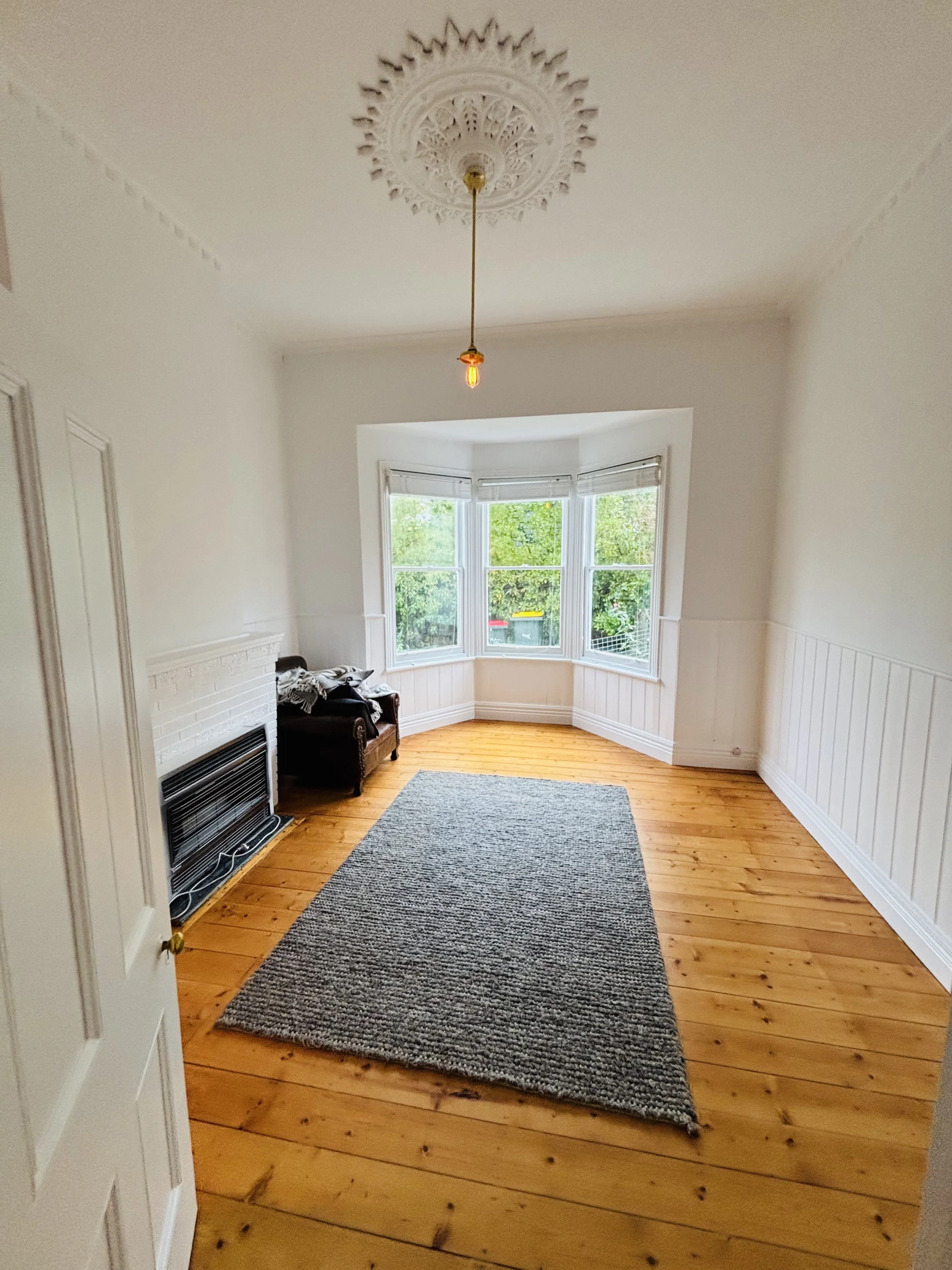 Empty living room with hardwood floors, a gray rug, a white brick fireplace, large bay window with greenery outside, and a single hanging light fixture.