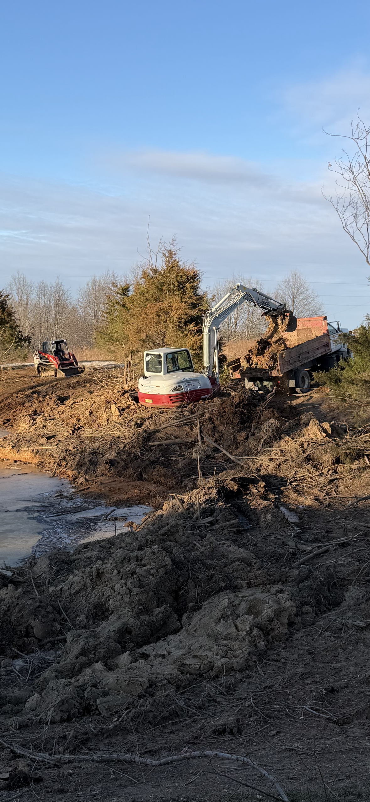 Excavator and truck clearing debris on a muddy shore near a body of water, with a forklift in the background and mostly leafless trees under a blue sky.