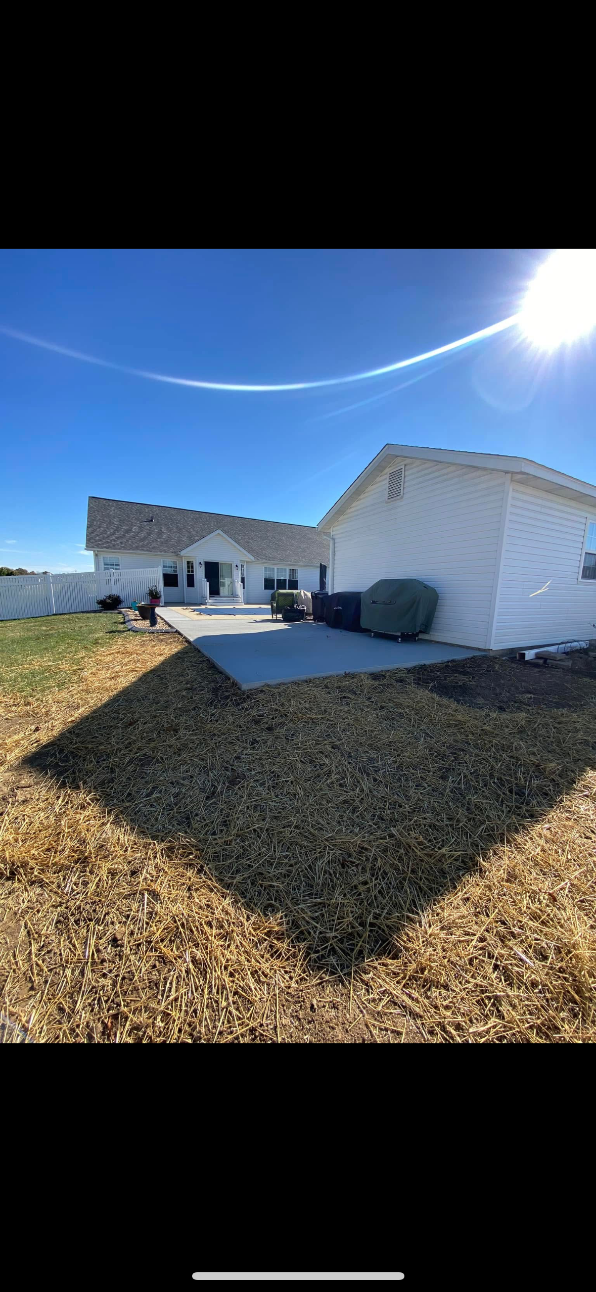 Backyard with a house, patio, grill covered with a green cover, and a white fence under a bright blue sky with the sun shining.