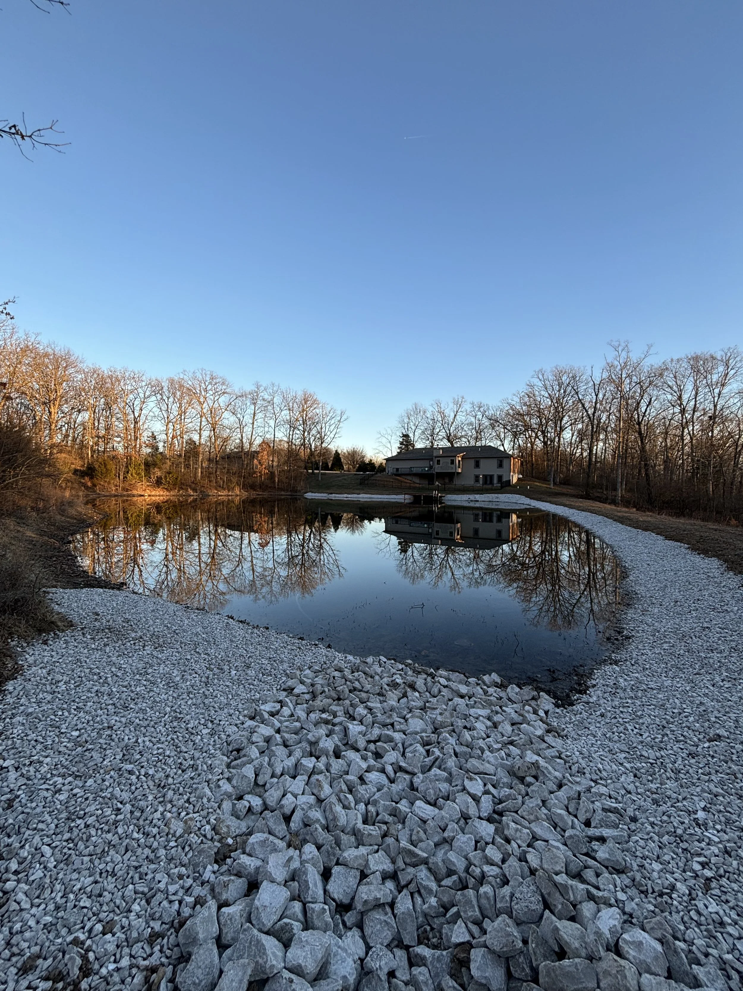 A calm river with a rocky bank on both sides, reflecting a house and leafless trees under a clear blue sky at sunset.
