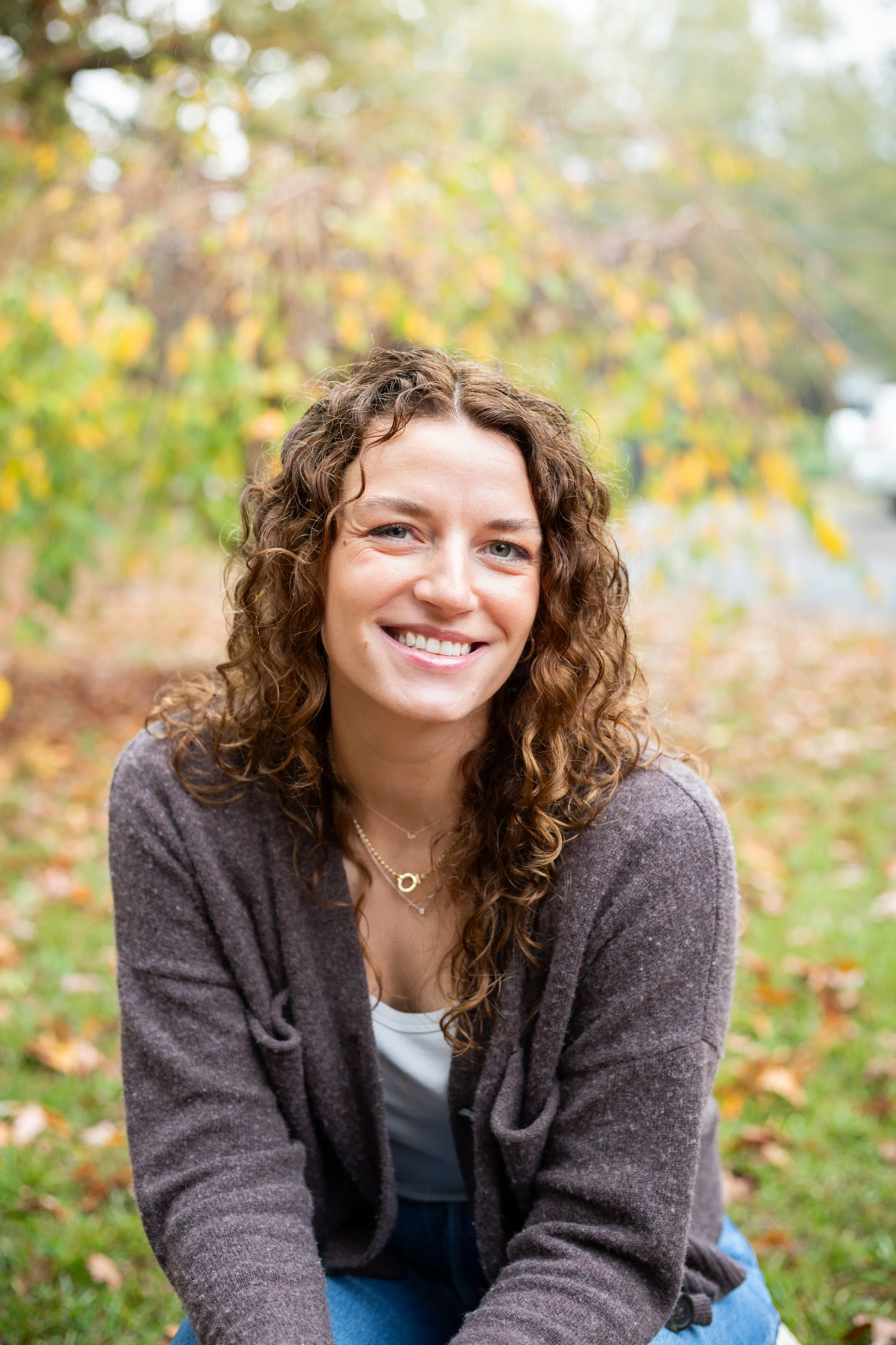 A woman with curly brown hair and a big smile, sitting outdoors in a park during fall with yellow and orange leaves in the background.
