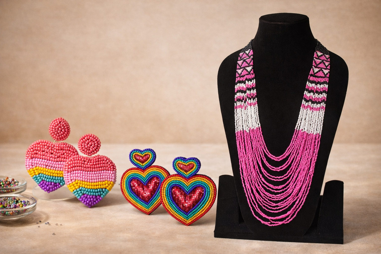 Display of colorful beaded jewelry, including layered pink and black necklace on a black bust, rainbow and heart-shaped earrings, and round earrings with rainbow stripes, alongside small bowls of sprinkles.