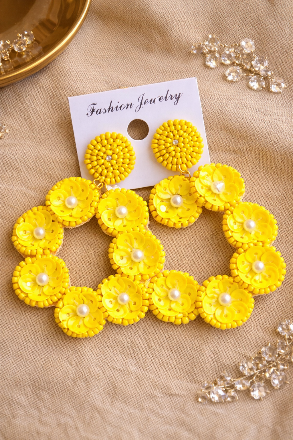 Yellow floral earrings with pearl centers displayed on a white card labeled "Fashion Jewelry" on a beige fabric surface, with additional crystal jewelry pieces nearby.