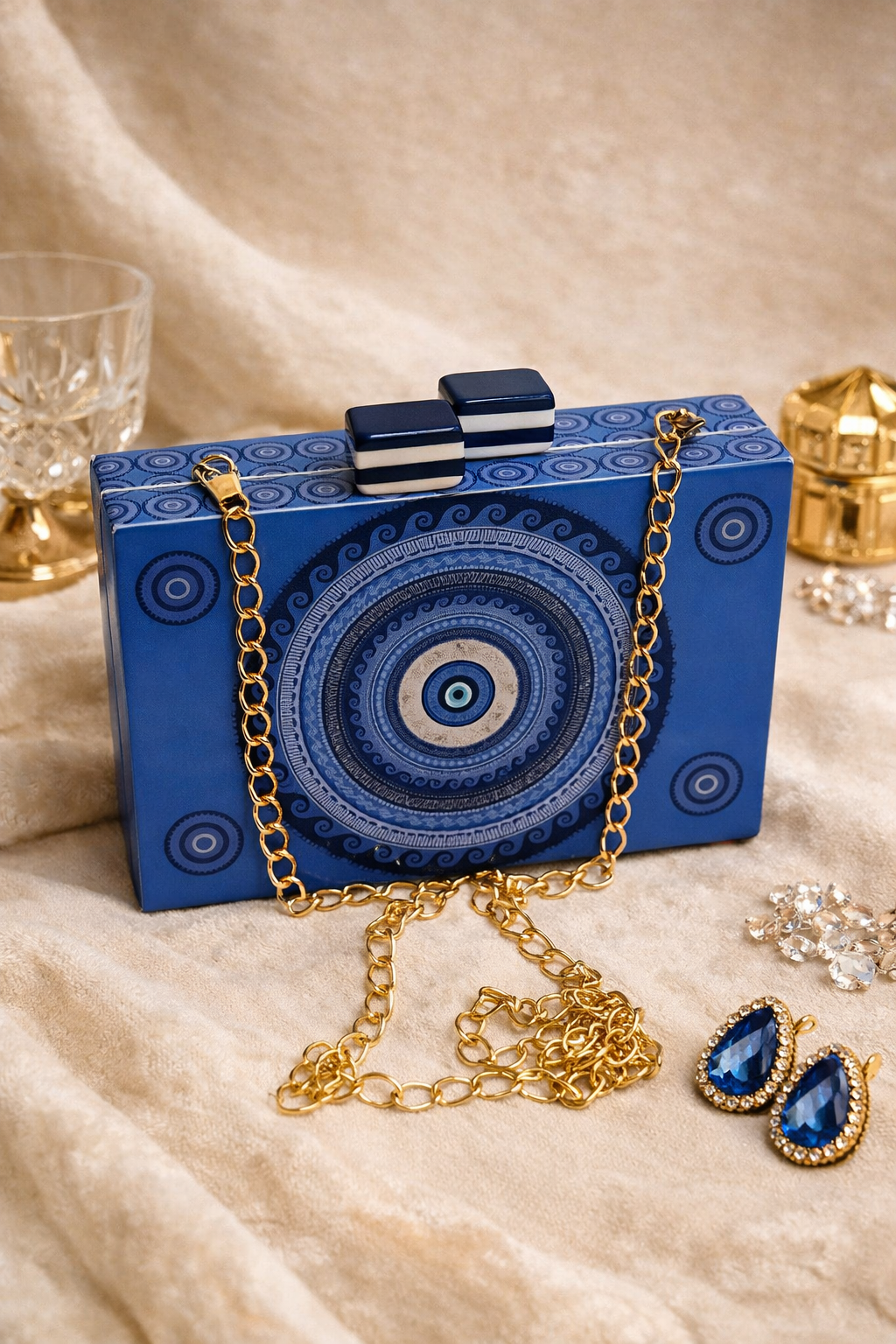 A decorative blue box with a circular pattern, adorned with a gold chain necklace and matching earrings, set on a beige fabric surface with crystal accents and glassware in the background.