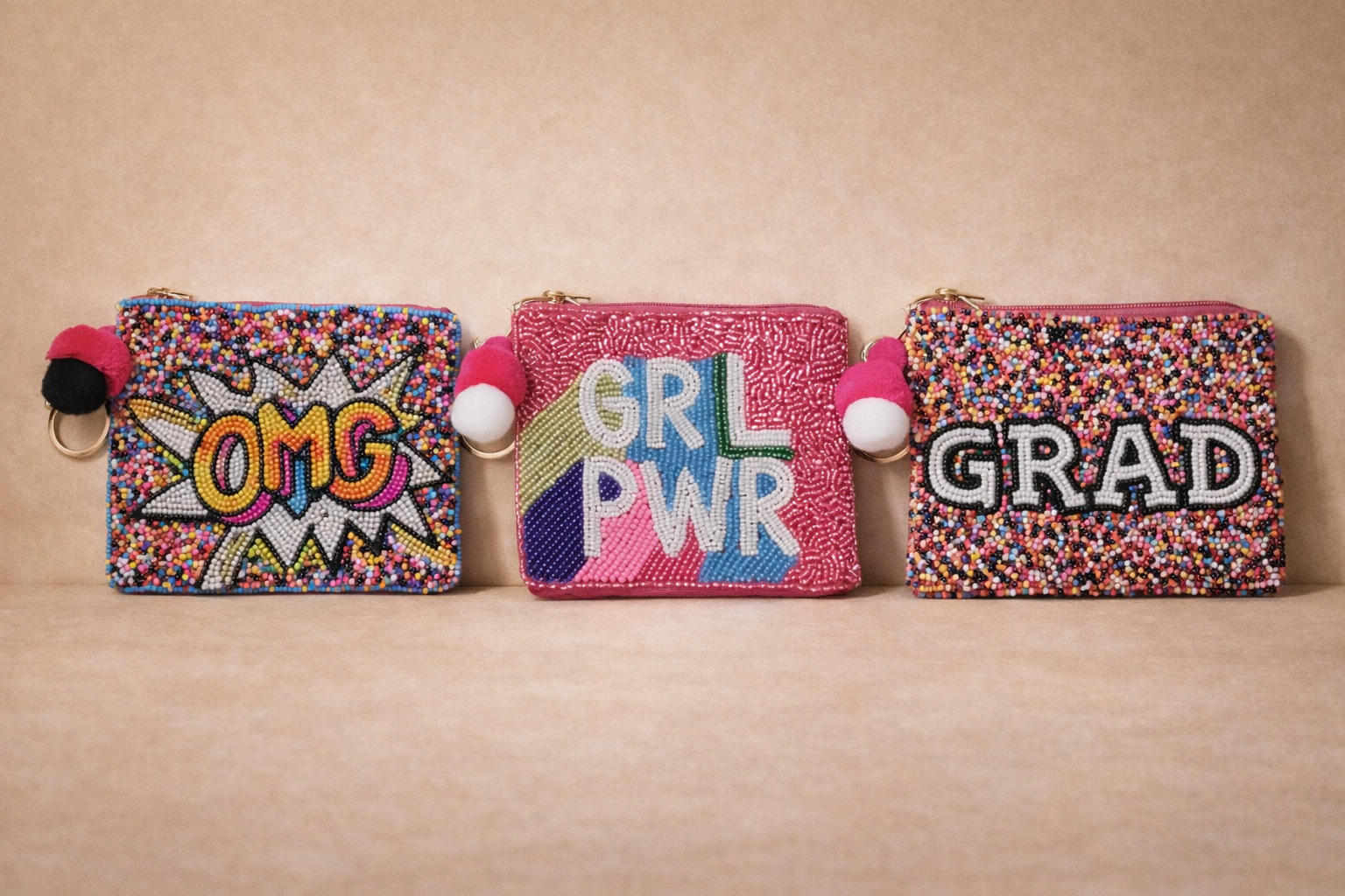 Three small beaded clutch purses with colorful designs and pom-pom keychains, arranged in a row on a beige surface. The first purse has 'OMG' in a comic style with multicolored beads, the middle purse features 'GRL PWR' with rainbow colors, and the t