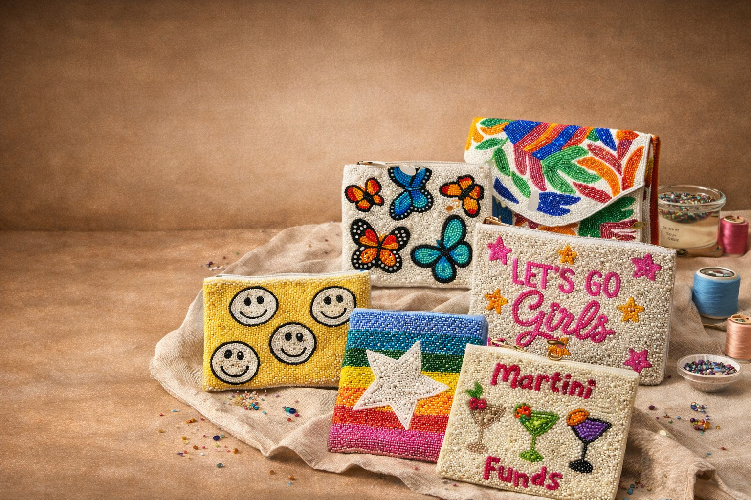 Colorful beaded bags with various designs, including butterflies, smiley faces, a rainbow with a star, and text such as "Let's go girls" and "Martini funds," arranged on a beige cloth with scattered beads and spools of thread in the background.