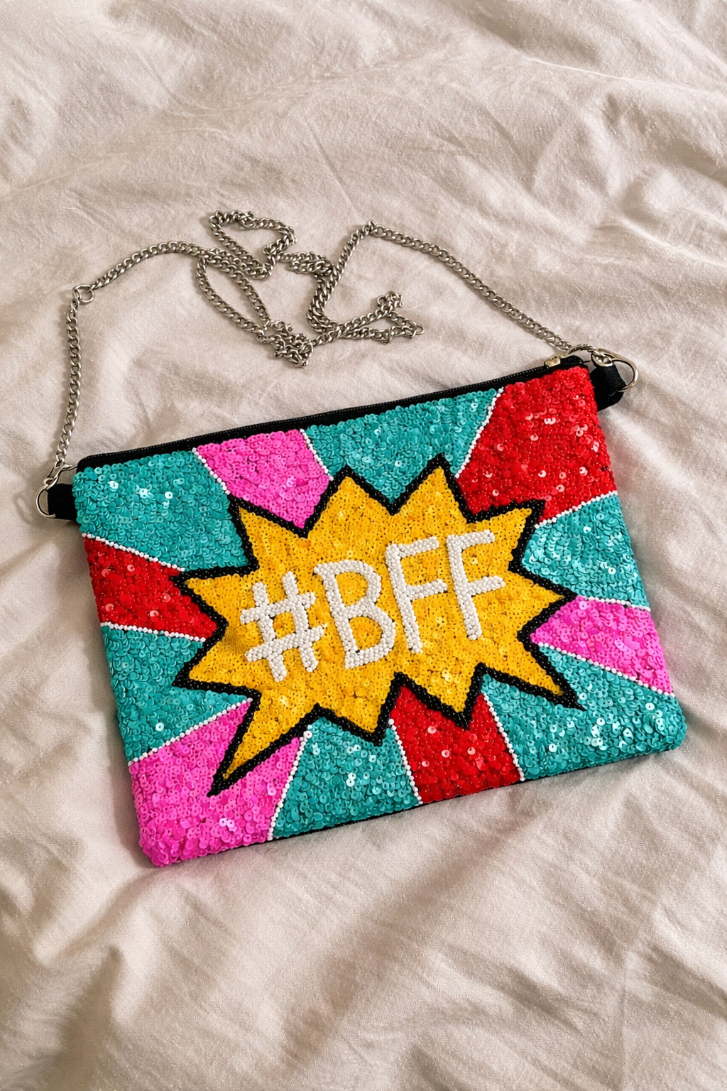 A colorful sequined clutch purse with a comic book style design that says '#BFF' in the center, lying on a soft, light-colored surface.