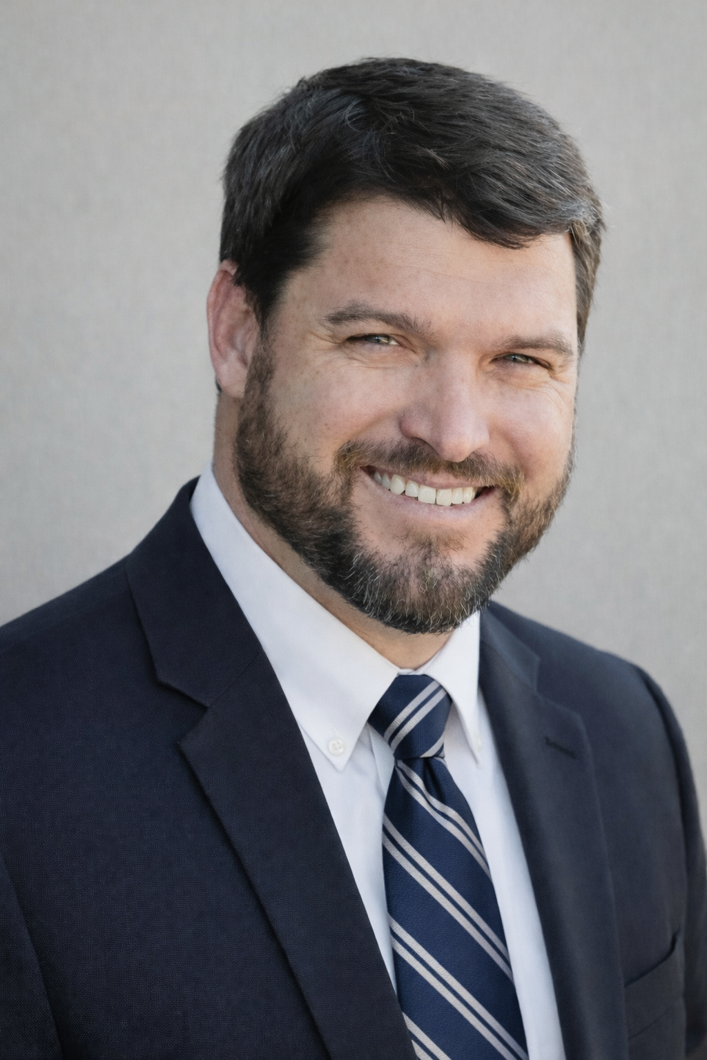 Professional headshot of a smiling man with dark hair and beard, wearing a navy suit, white shirt, and striped tie, against a plain gray background.