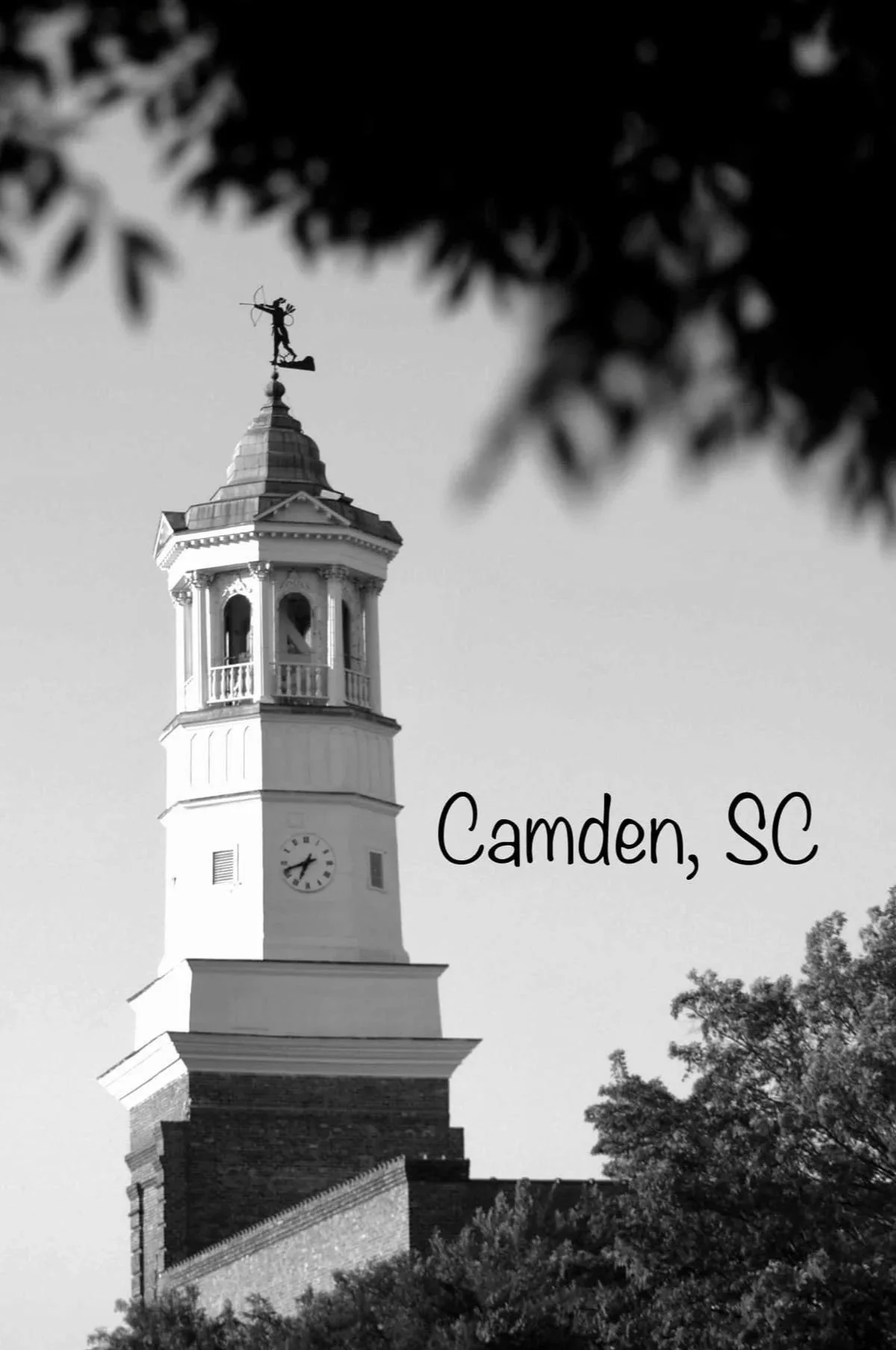 Black and white photo of a tall clock tower with a weathervane on top, surrounded by trees, with the words "Camden, SC" written at the lower right corner.