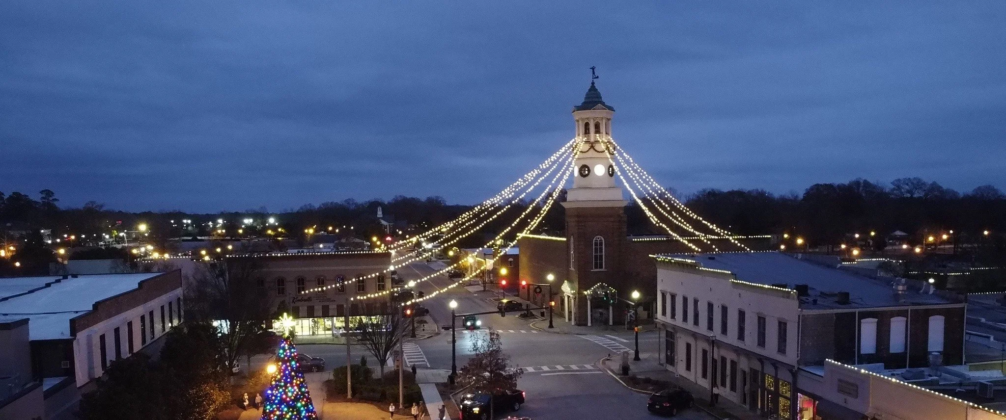 Nighttime view of a town square decorated with Christmas lights, featuring a tall clock tower with lights cascading down from the top, and a lit Christmas tree in the foreground.