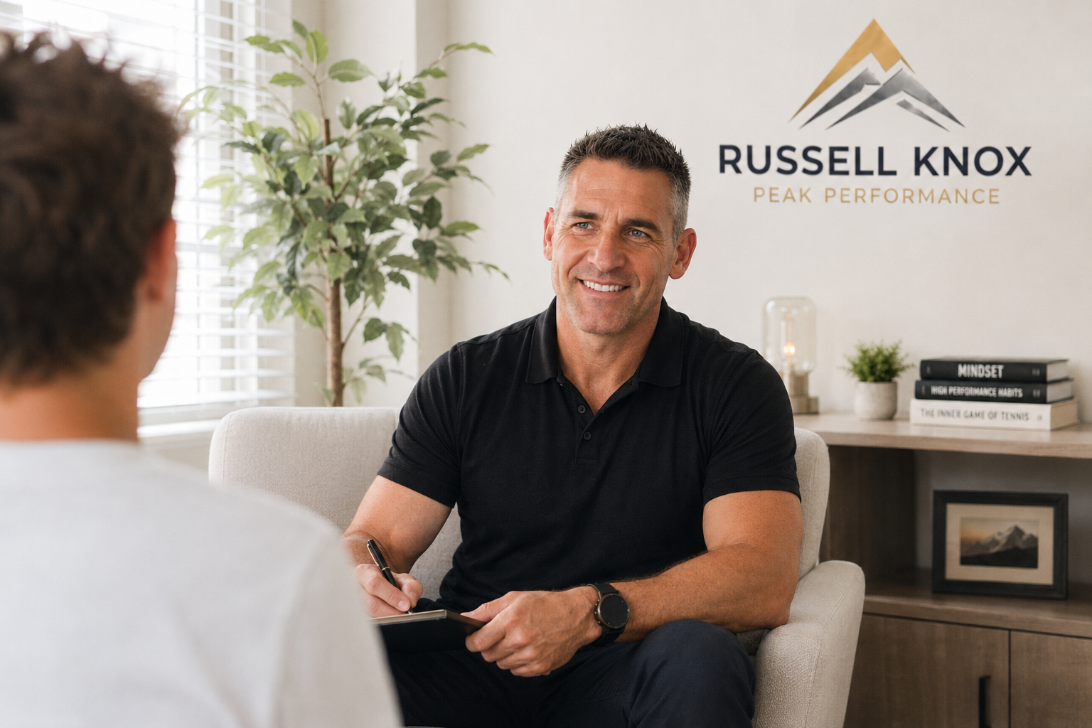 A man in a black shirt smiling during a consultation in an office with a Russell Knox Peak Performance sign on the wall.