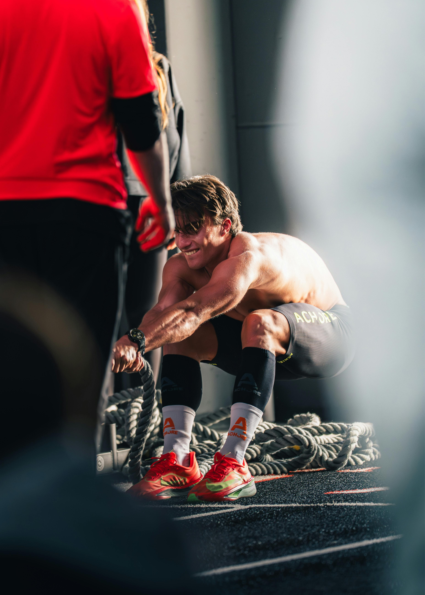 A shirtless man wearing black athletic shorts, white socks with orange and gray accents, and red Nike sneakers, crouching on a gym floor, smiling, with a person in red and black clothing holding a rope nearby.