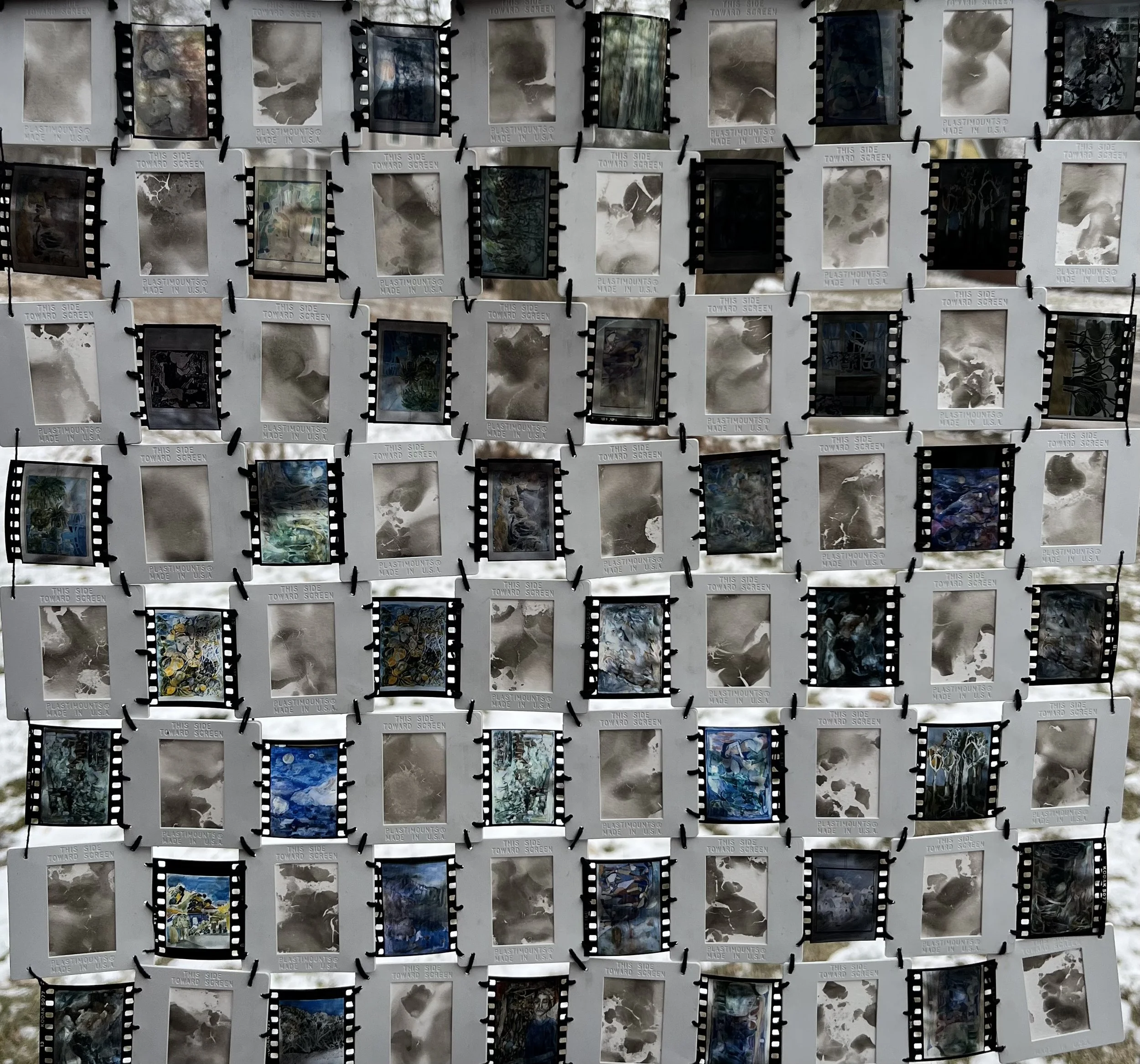 A collage of many small slides or film strips arranged in a grid, some with abstract patterns, others with scenes or textures, mounted on a display board outdoors.