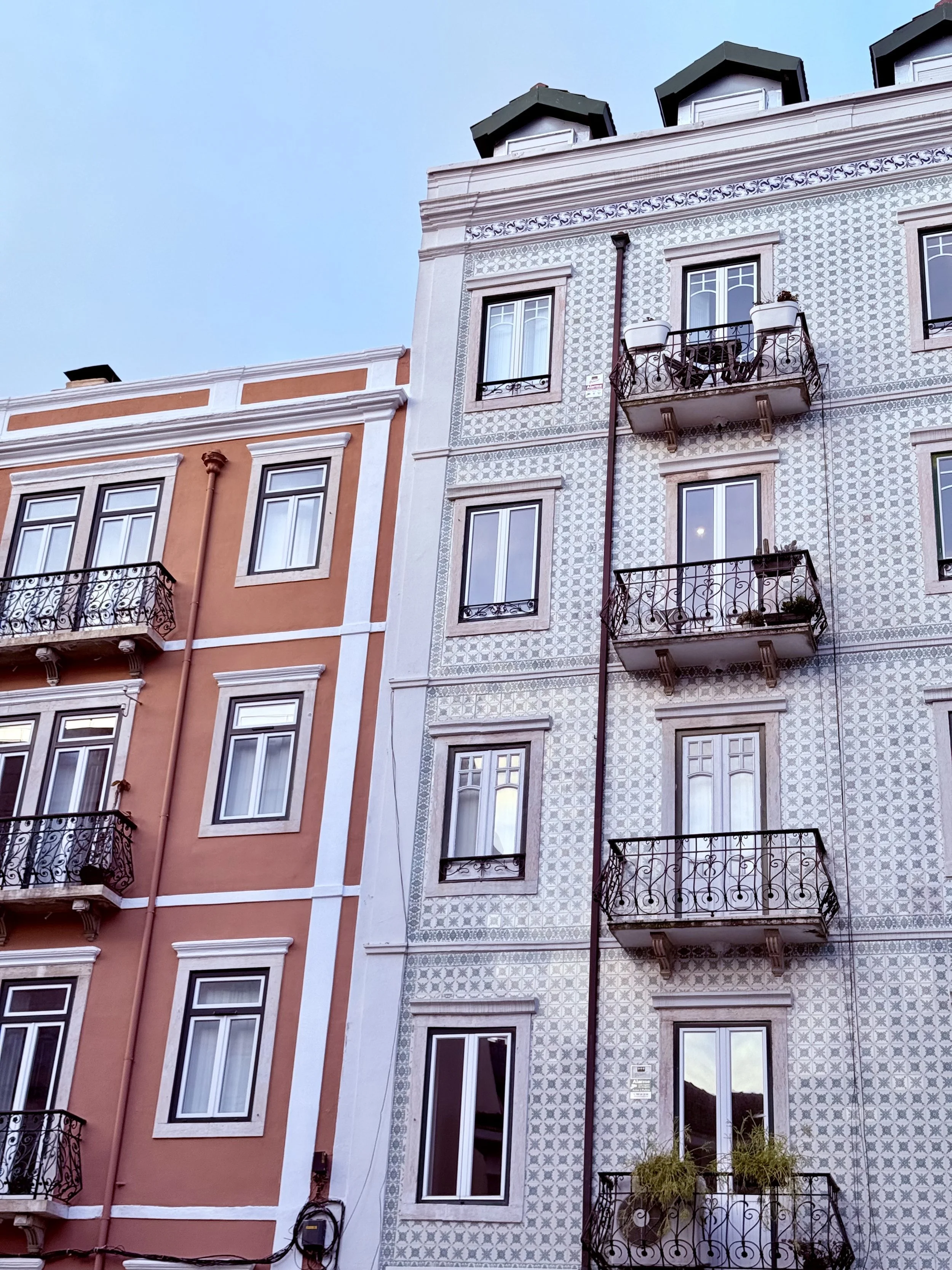 Close-up of two colorful apartment building facades, one in warm orange with black wrought-iron balconies and the other in white with detailed patterned tilework and matching balconies, under a clear sky.
