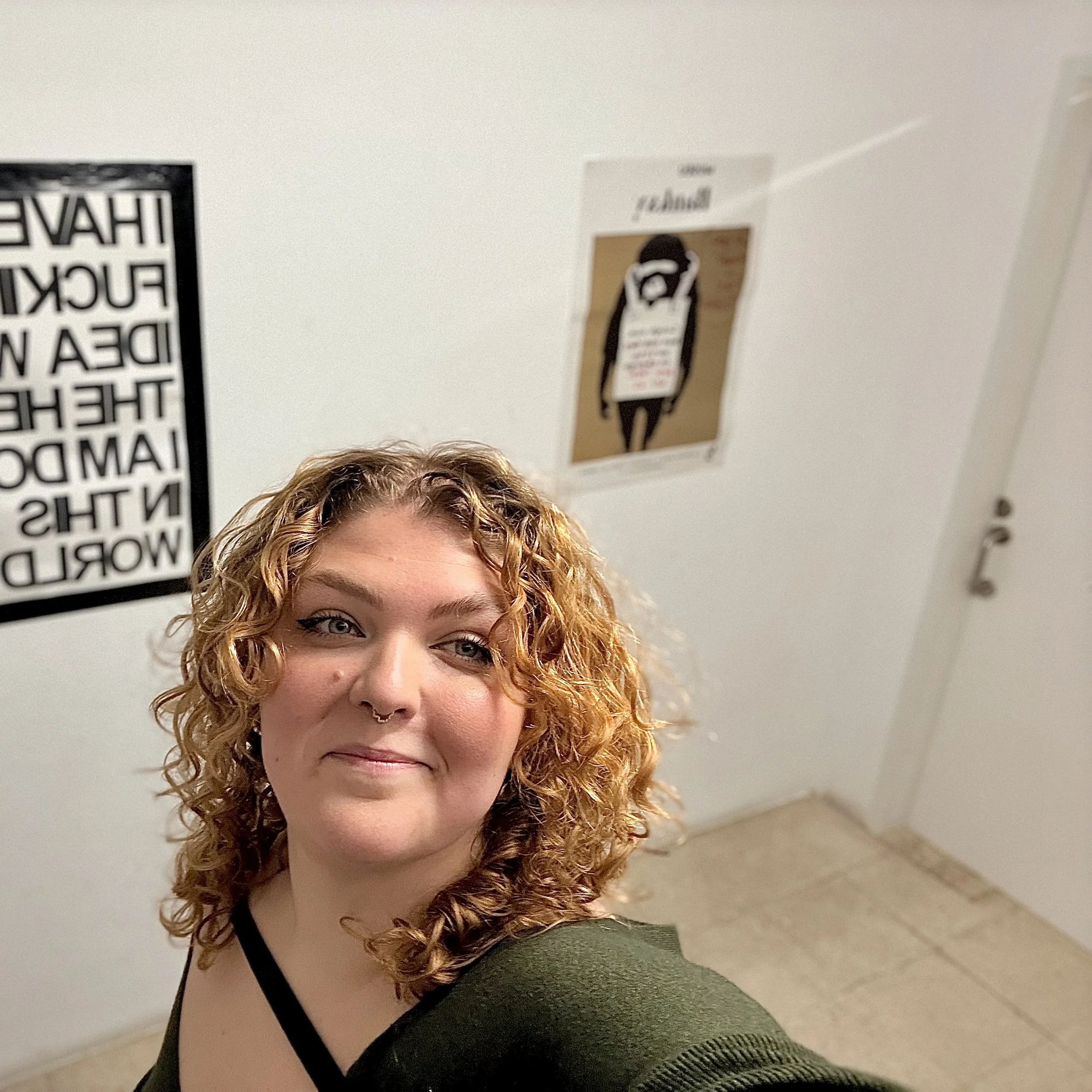 A woman with curly brown-blonde hair taking a selfie in an indoor setting, with posters on the wall behind her.