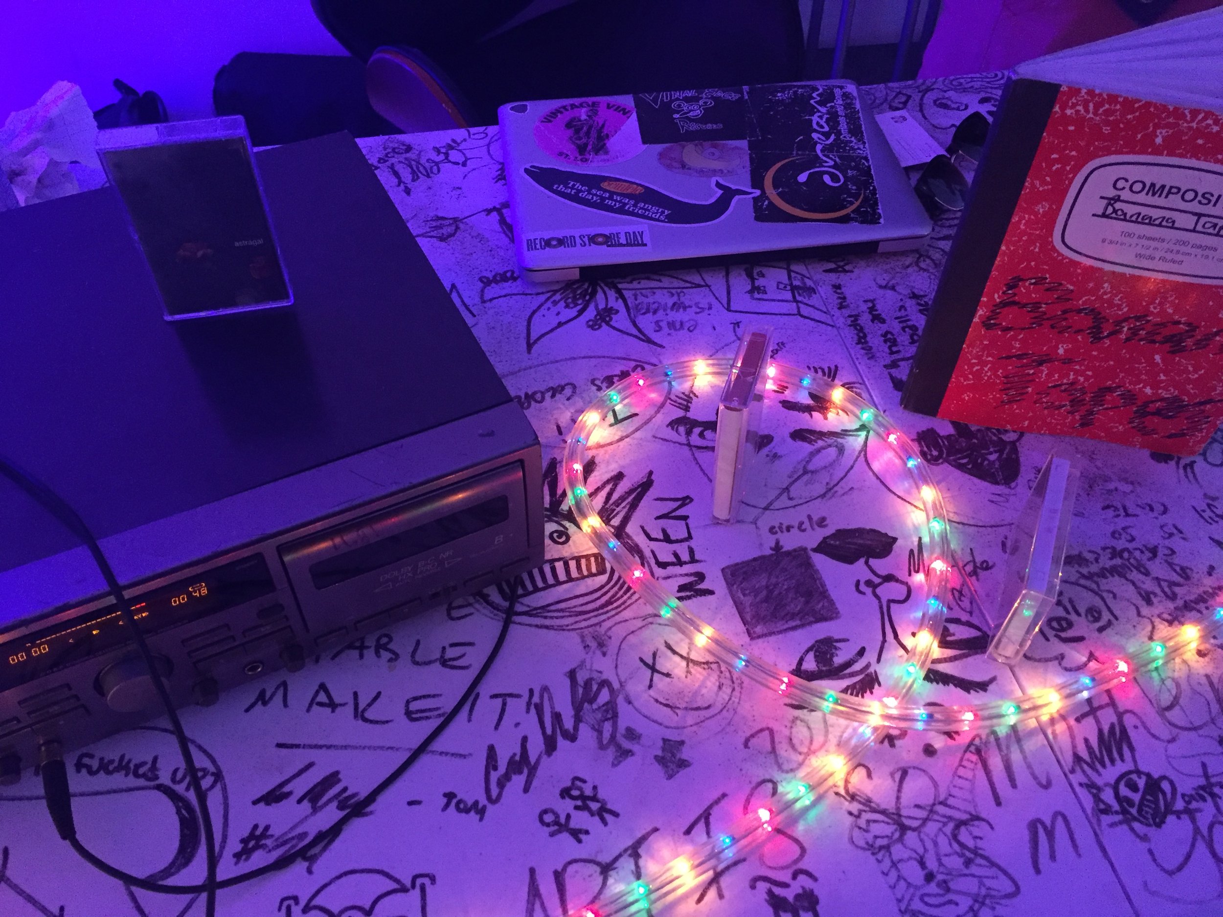 Table with drawing, a vintage stereo, a laptop with stickers, a box of composition notebooks, and colorful LED string lights in a dimly lit room.