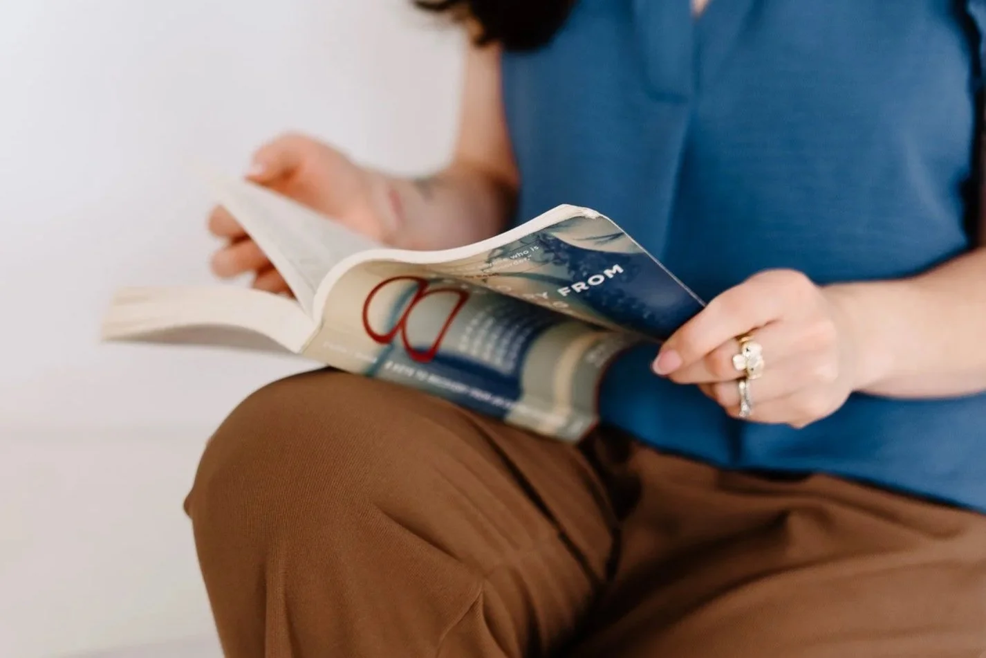 Person reading a magazine while sitting down, wearing a blue shirt and brown pants, with rings on their fingers.