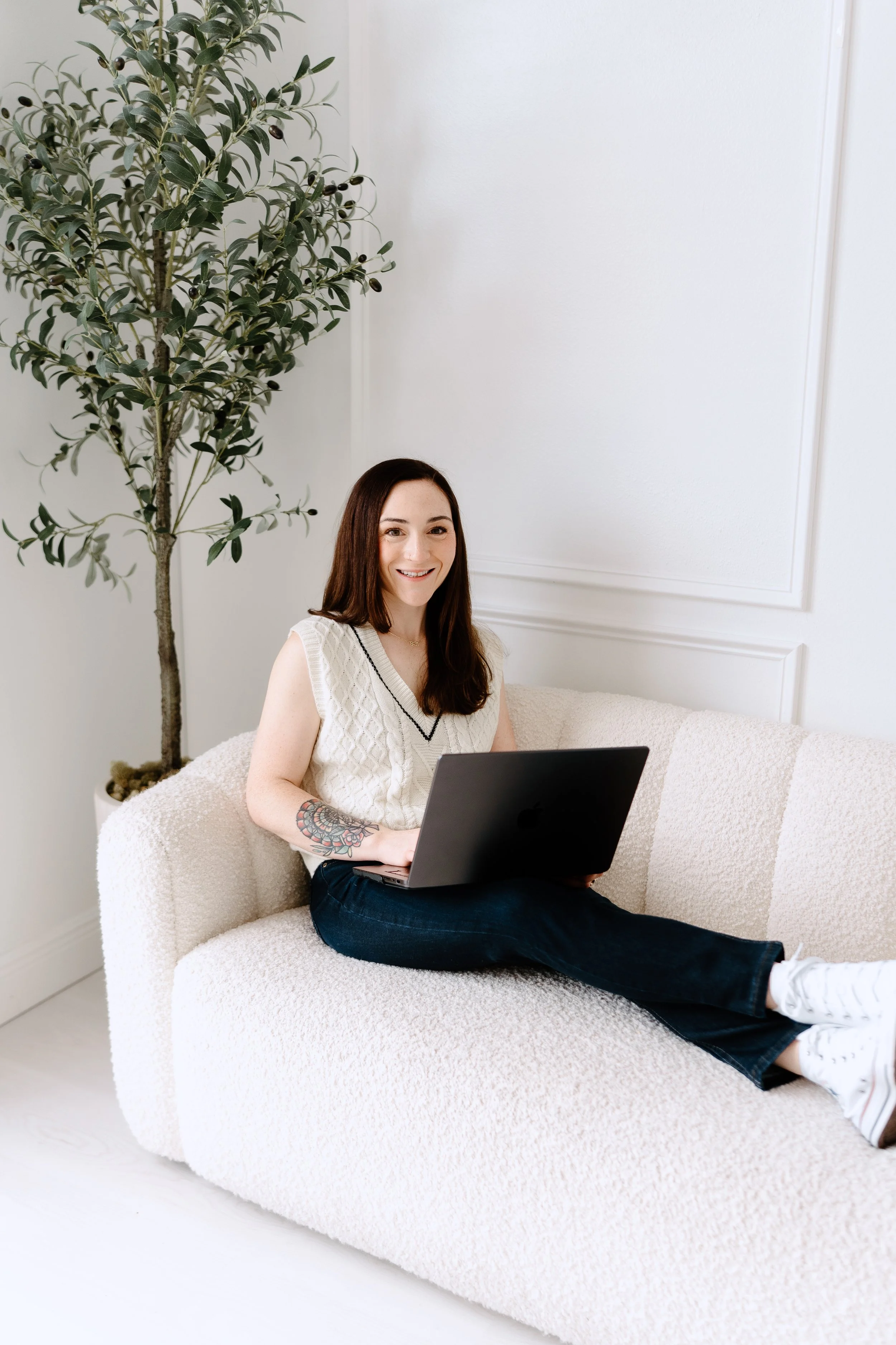 A young woman with long dark hair, wearing a white sleeveless sweater and jeans, sitting on a cream-colored sofa with a laptop on her lap. She has tattoos on her left forearm and is smiling at the camera. There is a tall potted plant with dark green leaves and black berries behind her, set against a white wall with decorative molding.