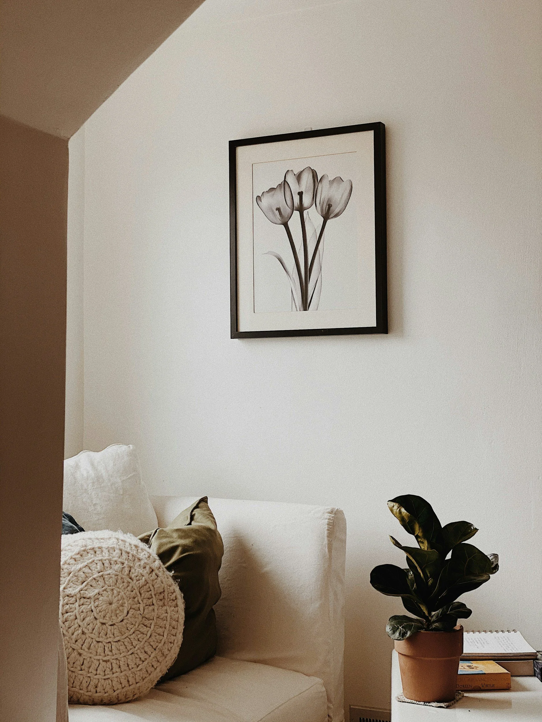 A cozy corner of a living room with a beige couch, layered with textured pillows. On the wall above, there is a framed black and white floral artwork. To the right, a potted plant with dark green, glossy leaves sits on a side table next to a few books and a calendar.