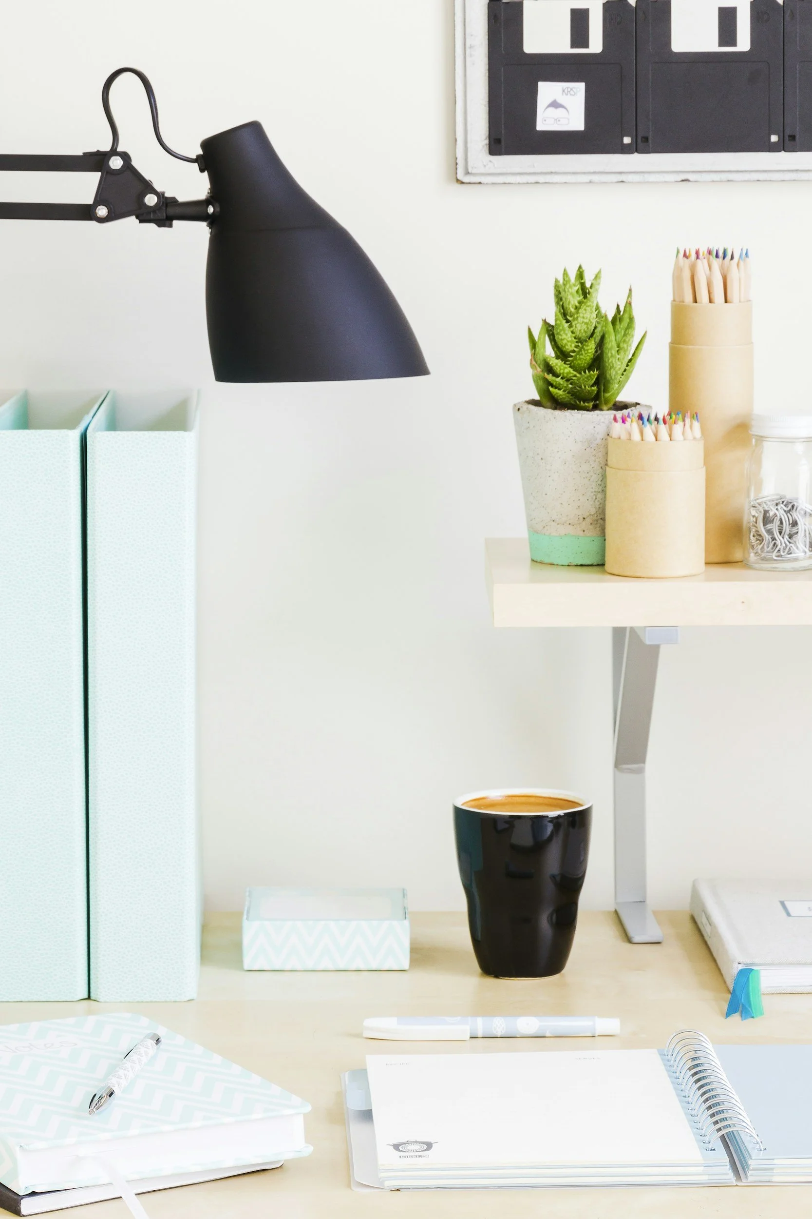 Office desk with a black desk lamp, a potted succulent plant in a concrete pot, two cardboard containers with colored pencils, a glass jar filled with paperclips, and a coffee mug.