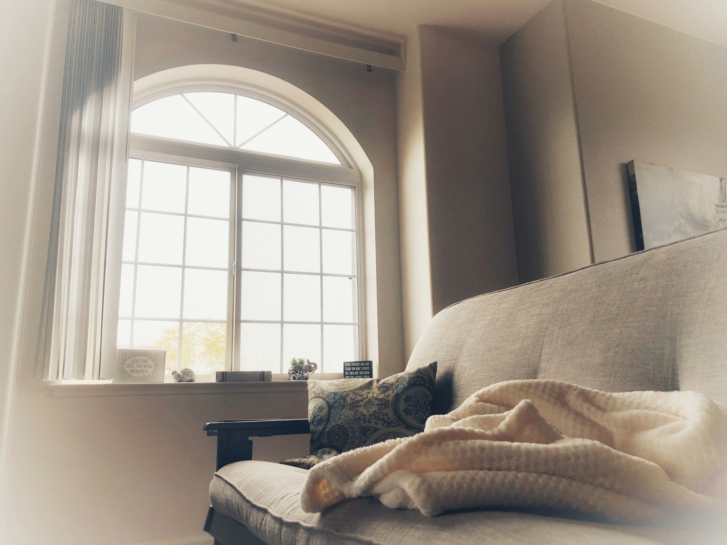 A cozy living room with a large arched window, a beige sofa with decorative pillows, a fluffy cream blanket, and a small table displaying framed quotes, plants, and ornaments.