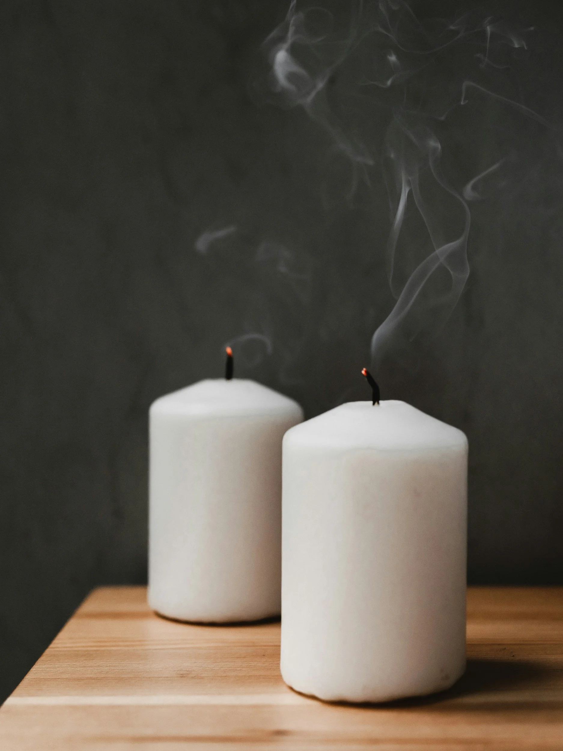 Two white candles on a wooden surface with thin smoke rising from burnt wicks, set against a dark background.