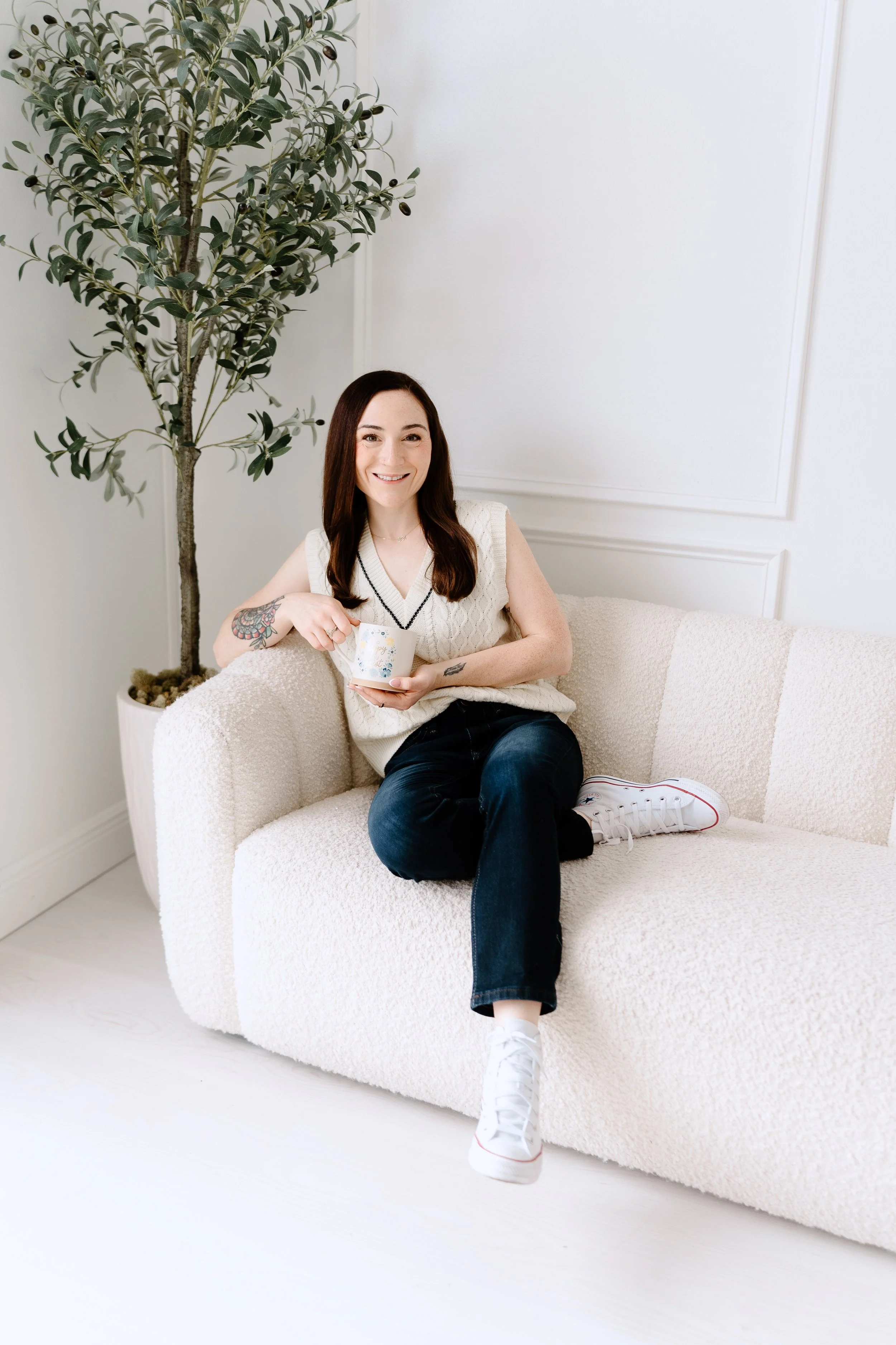 A woman with long dark hair, sitting on a cream-colored sofa, smiling, holding a mug, wearing a sleeveless cream sweater, jeans, and white sneakers, with a large potted plant behind her in a bright, minimalist room.