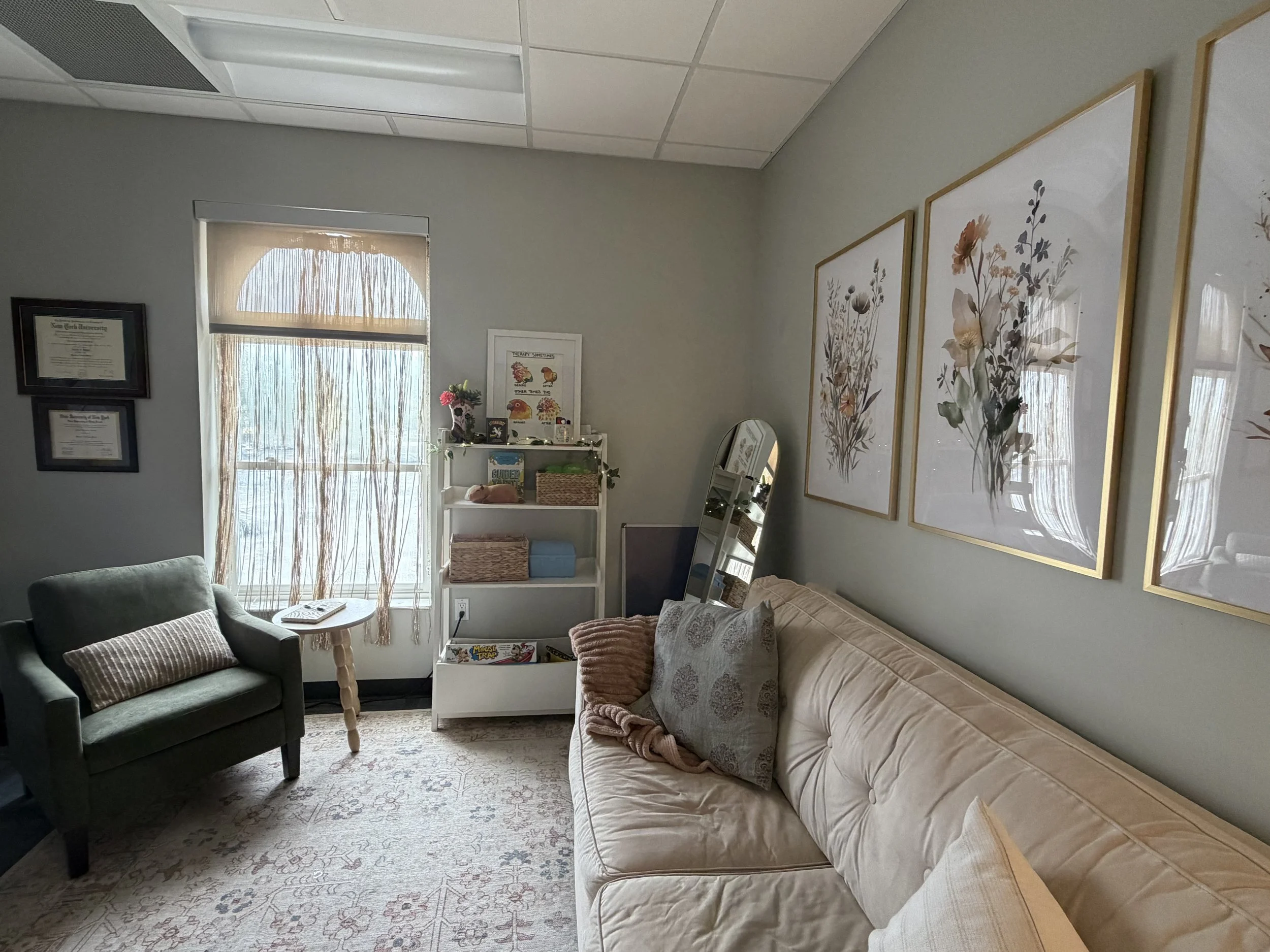 A cozy living room with a gray armchair, a beige sofa with pillows, a white bookshelf with decor and books, a small mirror, and framed botanical art on the wall. There is a window with sheer curtains, allowing natural light into the room, which has a patterned area rug on the floor.