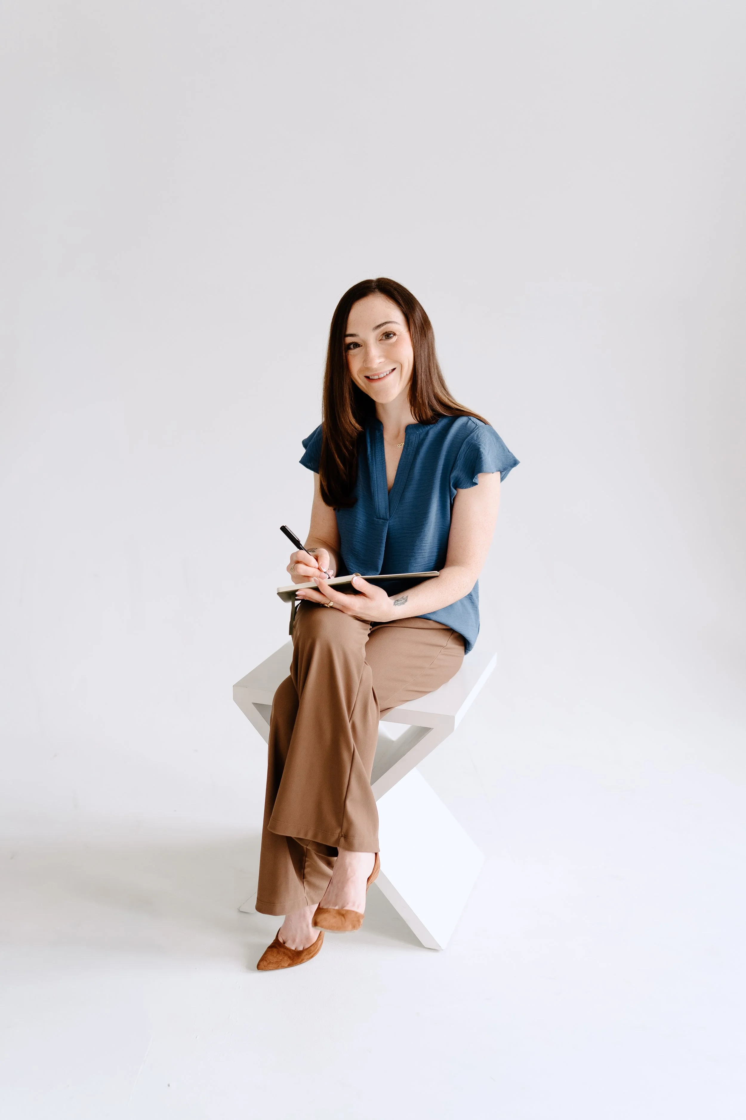 A woman with long brown hair, wearing a blue blouse, tan pants, and brown shoes, sitting on a modern white chair in front of a plain white background, smiling and holding a tablet and pen.
