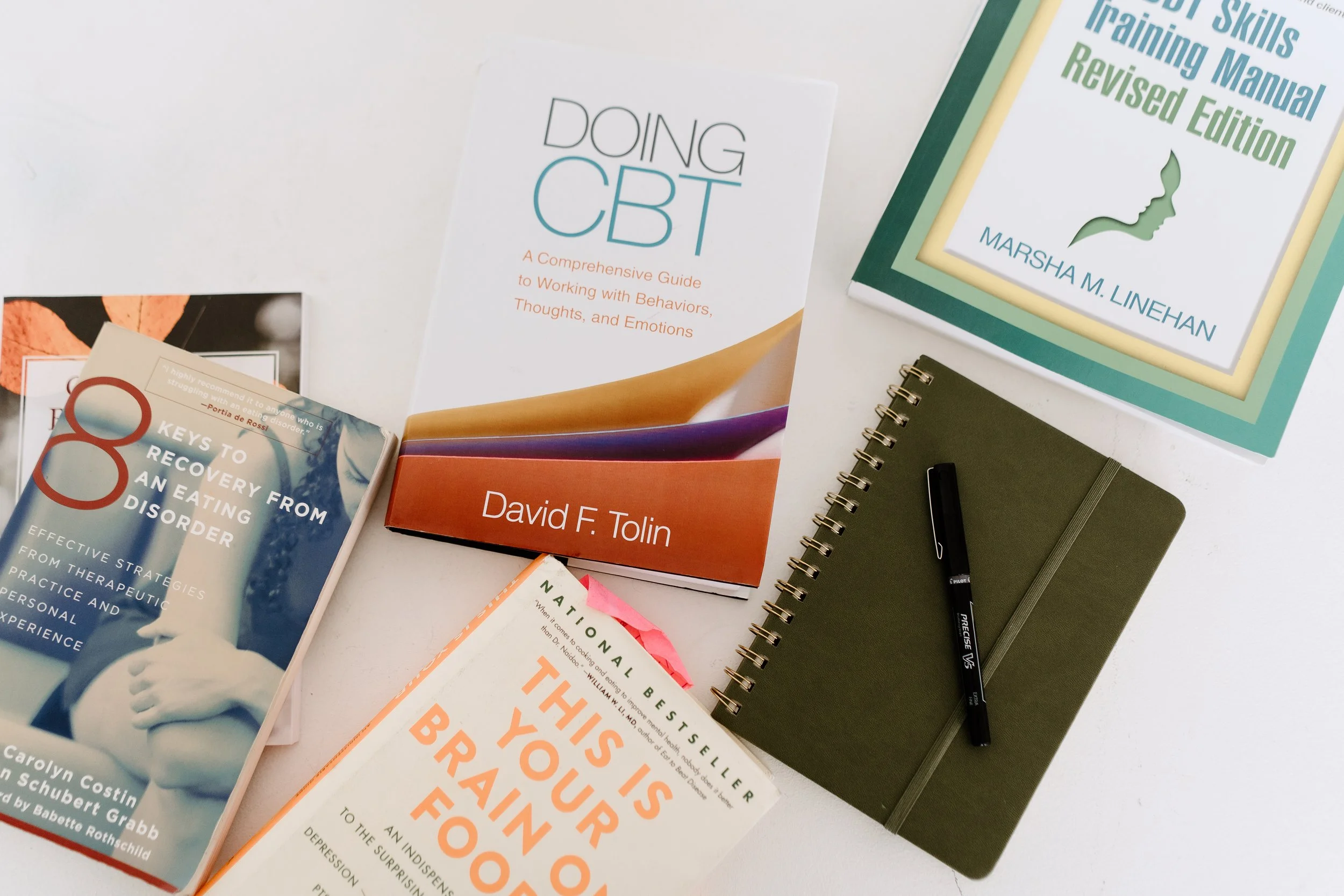 Table displaying various books and notebooks related to behavioral therapy and personal development, including titles like "Doing CBT", "Skills Training Manual Revised Edition", and "The House Is Burning" along with a black pen and a green notebook.