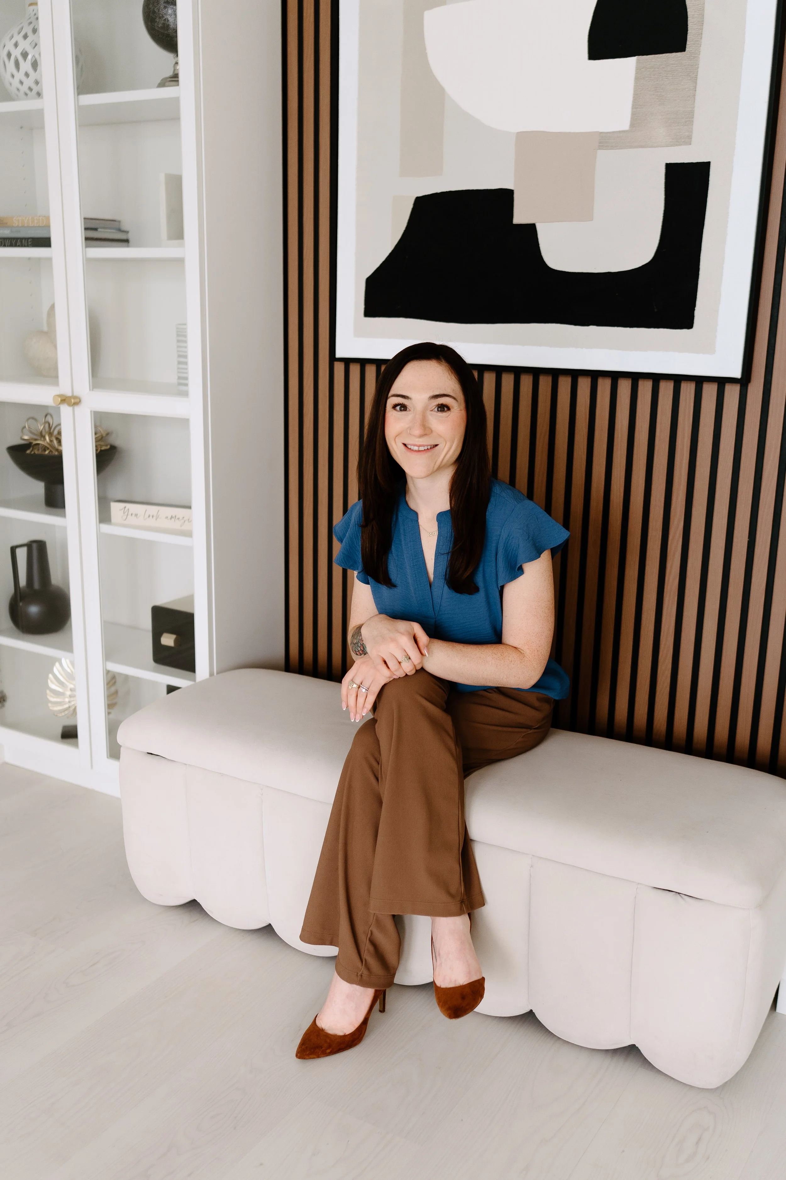A woman with long dark hair and a bright smile, sitting on a white upholstered bench, wearing a blue blouse, brown pants, and brown pointed-toe heels, with an abstract black, white, beige, and gray artwork hanging on a wooden-paneled wall behind her, and a white display cabinet with decorative objects to her left.