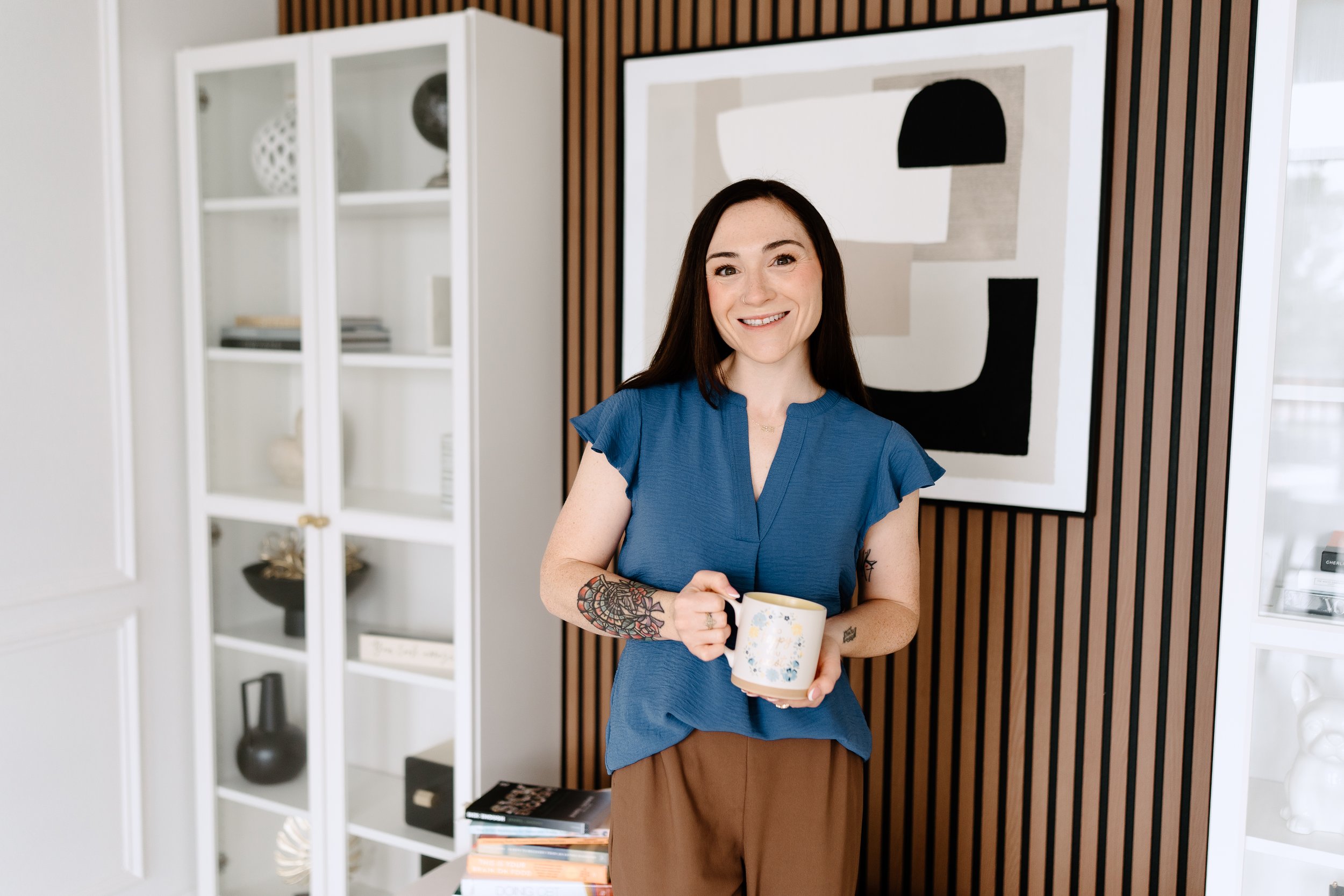 A woman with long dark hair and tattoos on her left arm, smiling and holding a coffee mug in a modern, well-lit room with white shelves and geometric wall art.