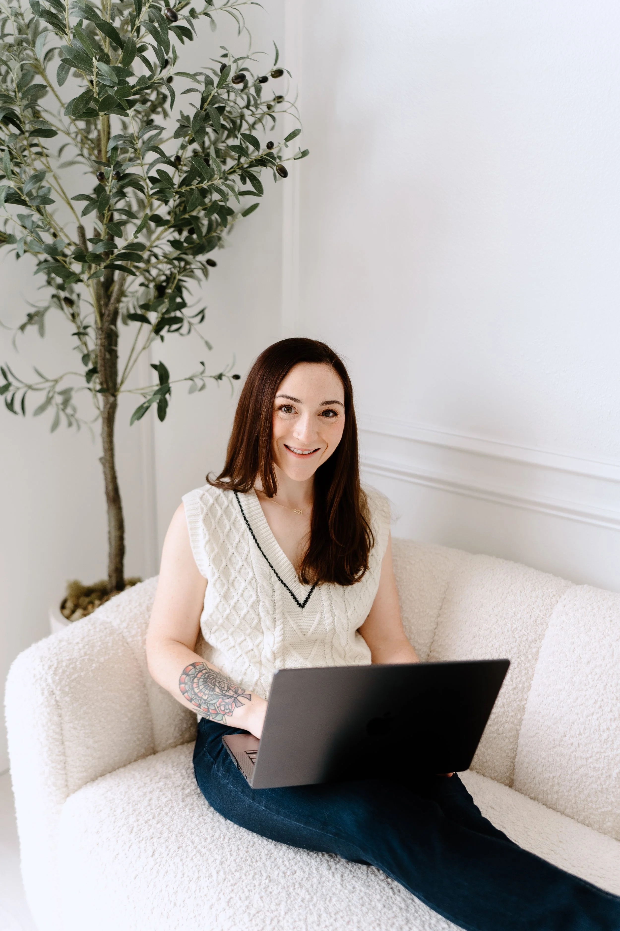 A woman with long brown hair, wearing a sleeveless white top with black trim, sitting on a cream-colored sofa, smiling and working on a laptop. She has a colorful tattoo on her left arm and is in a room with white walls and a tall indoor plant with green leaves.