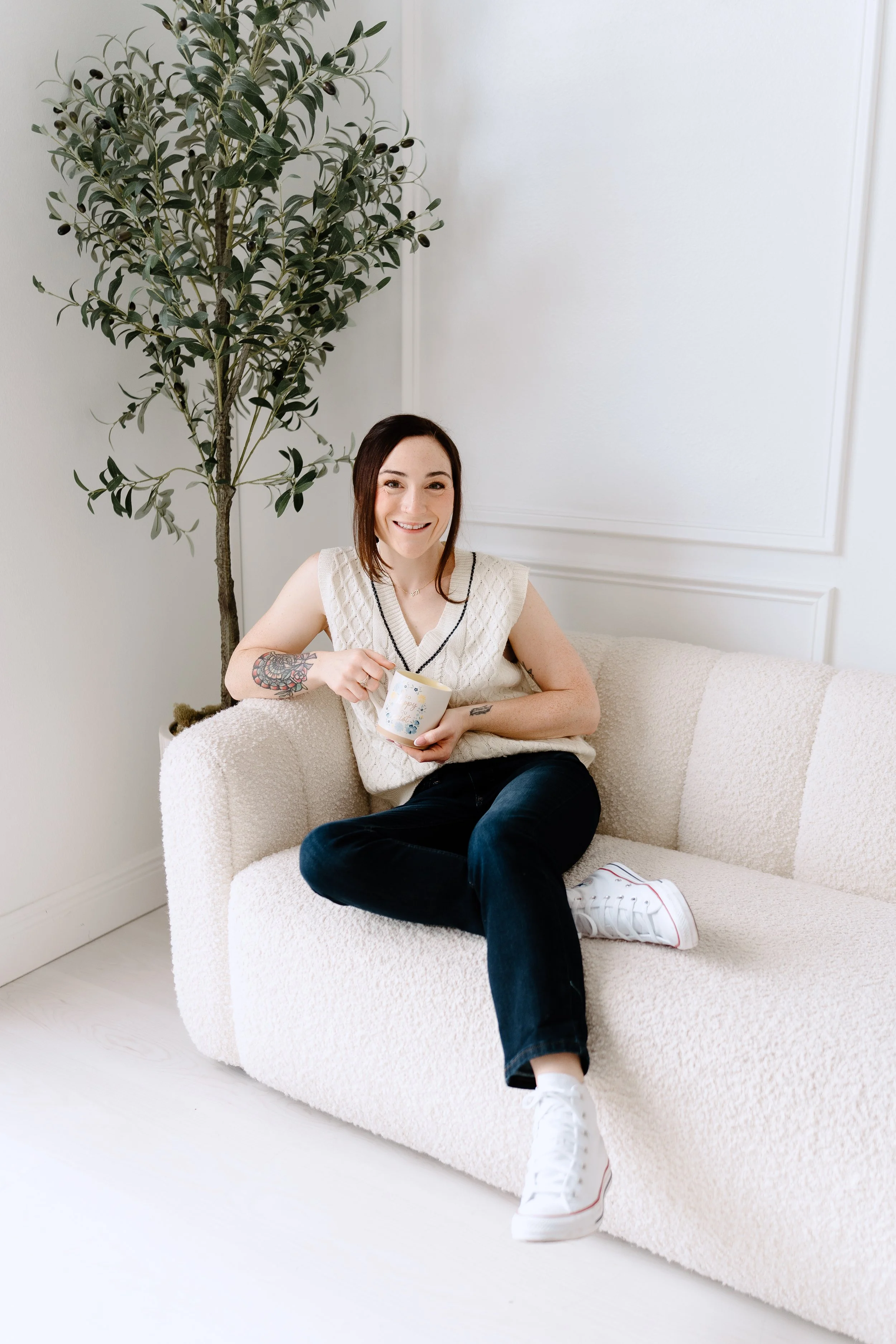 A young woman with tattoos on her left arm, smiling and sitting on a white textured sofa, holding a mug. She is wearing a sleeveless white knitted top, black pants, and white sneakers. There is a tall potted plant with dark green leaves and small black berries behind her, against a plain white wall with decorative paneling.