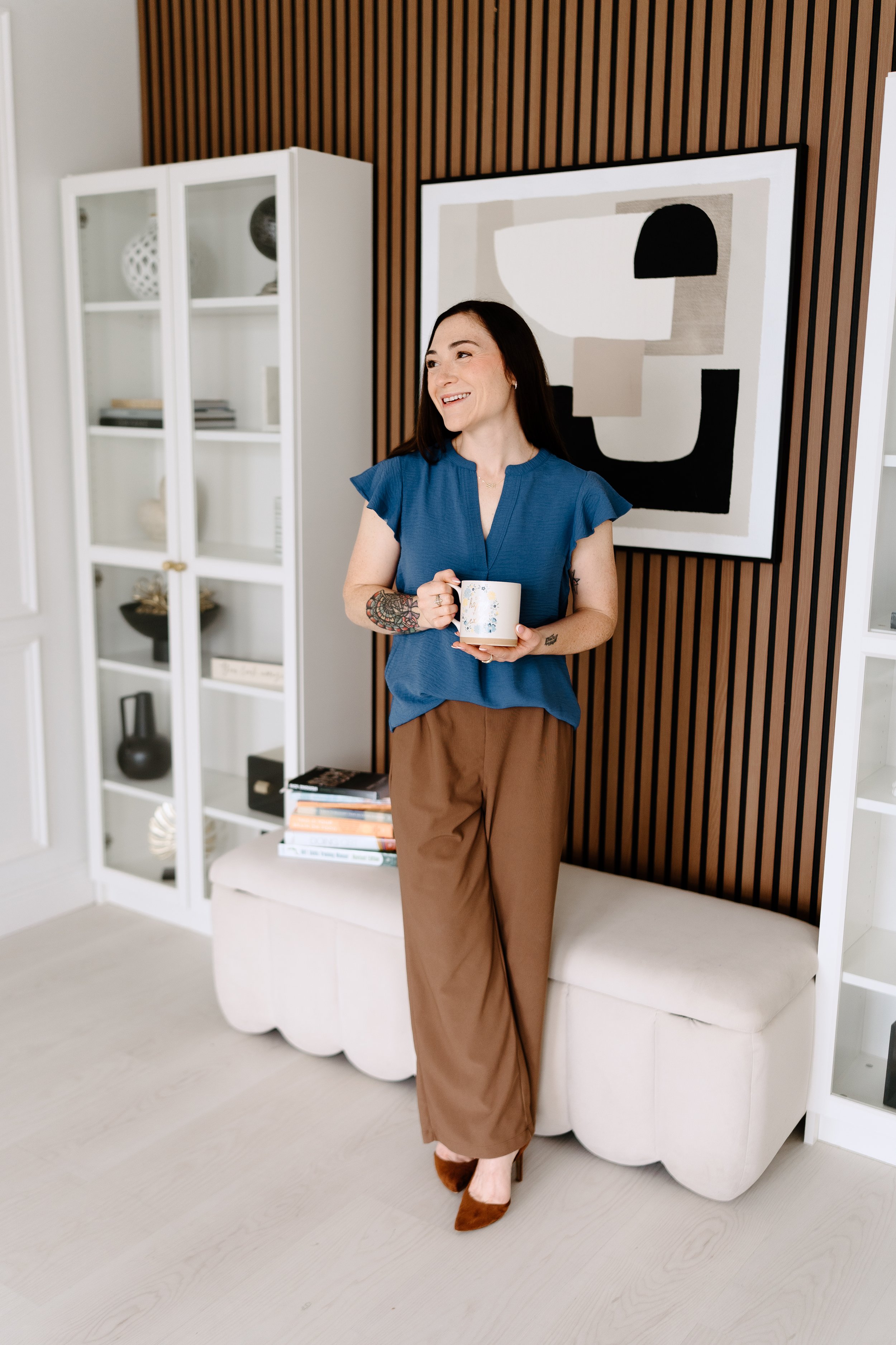 A woman standing in a modern living room, holding a coffee mug, smiling, wearing a blue top and brown pants.
